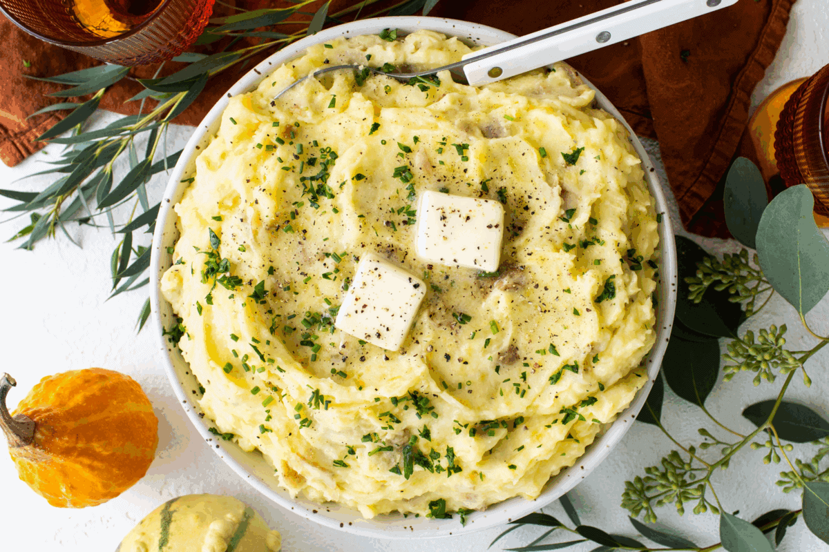 A bowl of mashed potatoes topped with two pats of butter, chopped herbs, and ground pepper sits amid autumn-themed decorations—one of the best Thanksgiving sides for your holiday table.