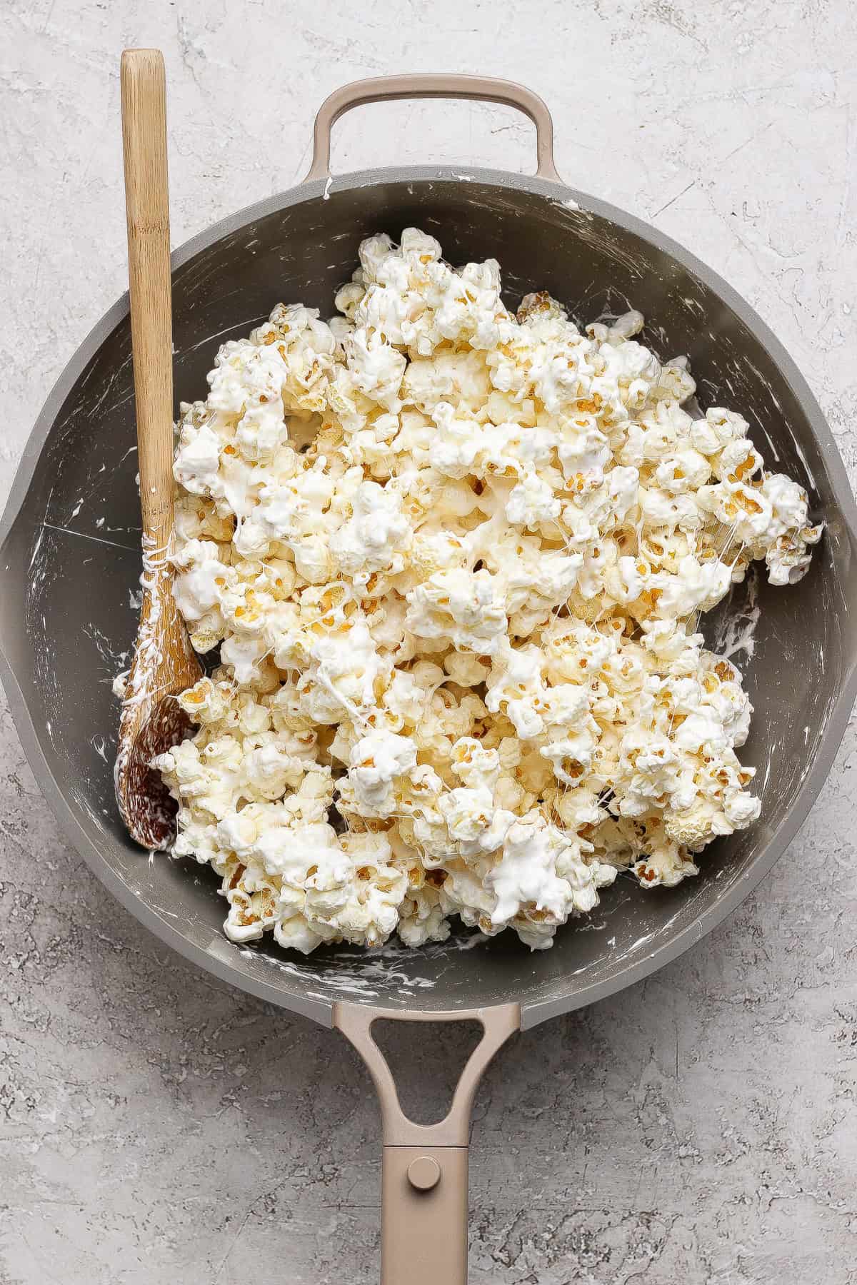 A pan filled with popcorn coated in a white, sticky mixture, with a wooden spoon resting on the side.
