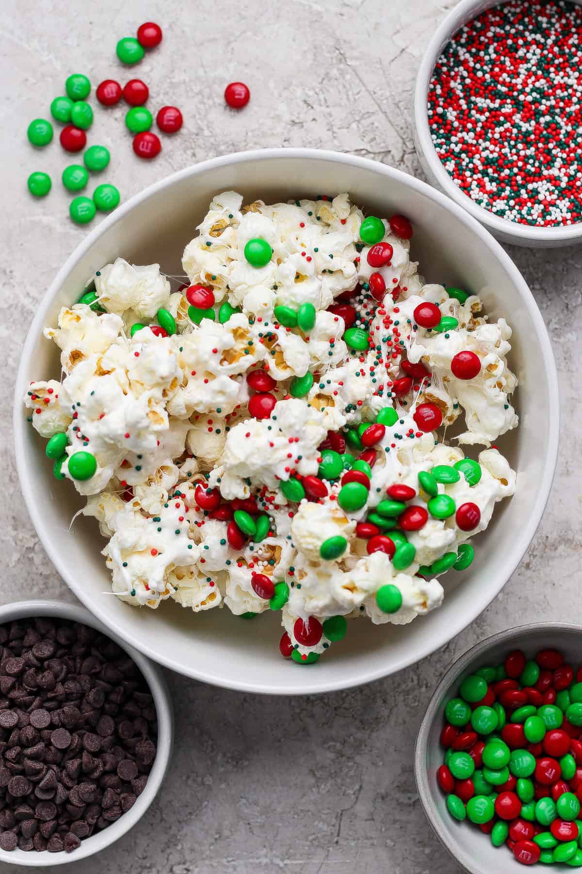 A bowl of popcorn topped with red and green candy-coated chocolates and sprinkles, surrounded by bowls of chocolate chips, more candies, and sprinkles on a light surface.