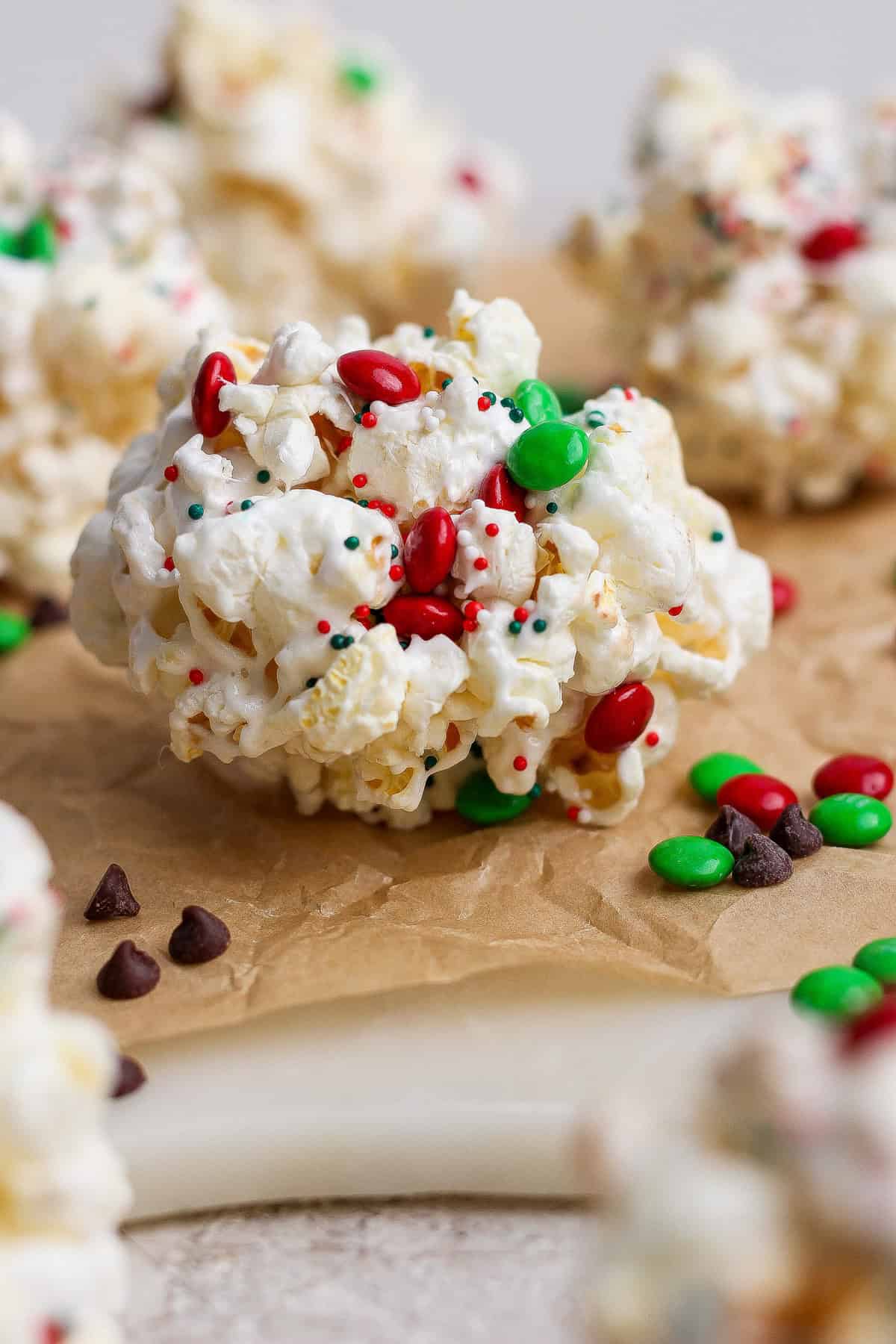 A close-up of a popcorn ball decorated with red and green candy-coated chocolates and multicolored sprinkles, placed on brown parchment paper.