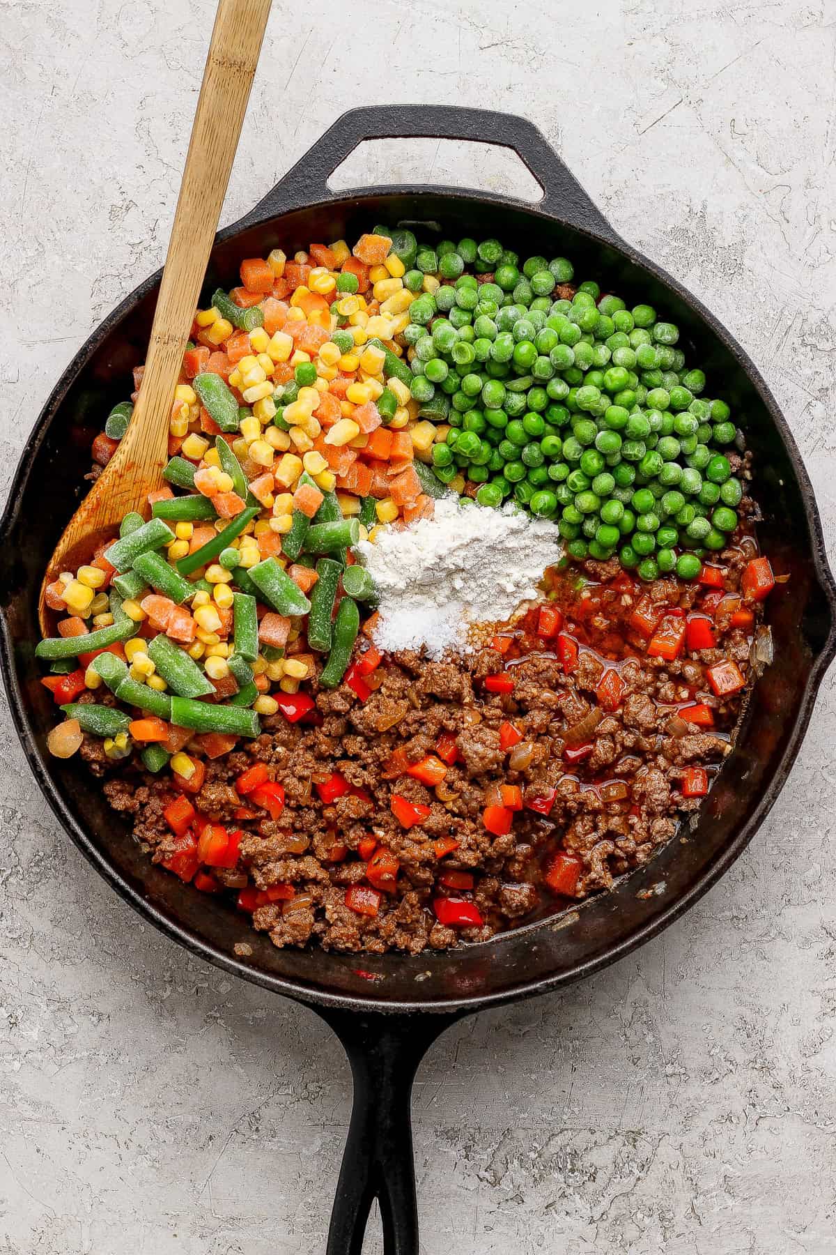 A cast iron skillet containing ground beef, peas, corn, green beans, diced red peppers, flour, and sauce, with a wooden spoon resting inside.