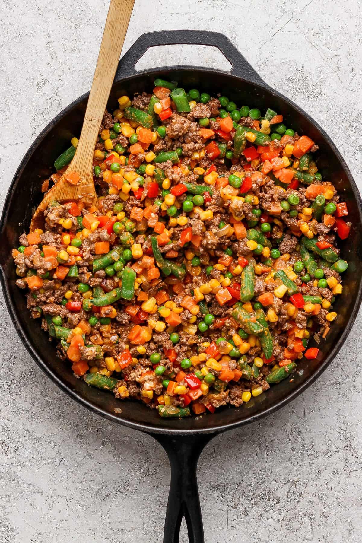 A cast iron skillet filled with cooked ground beef mixed with assorted vegetables, including peas, corn, carrots, and green beans, with a wooden spatula.