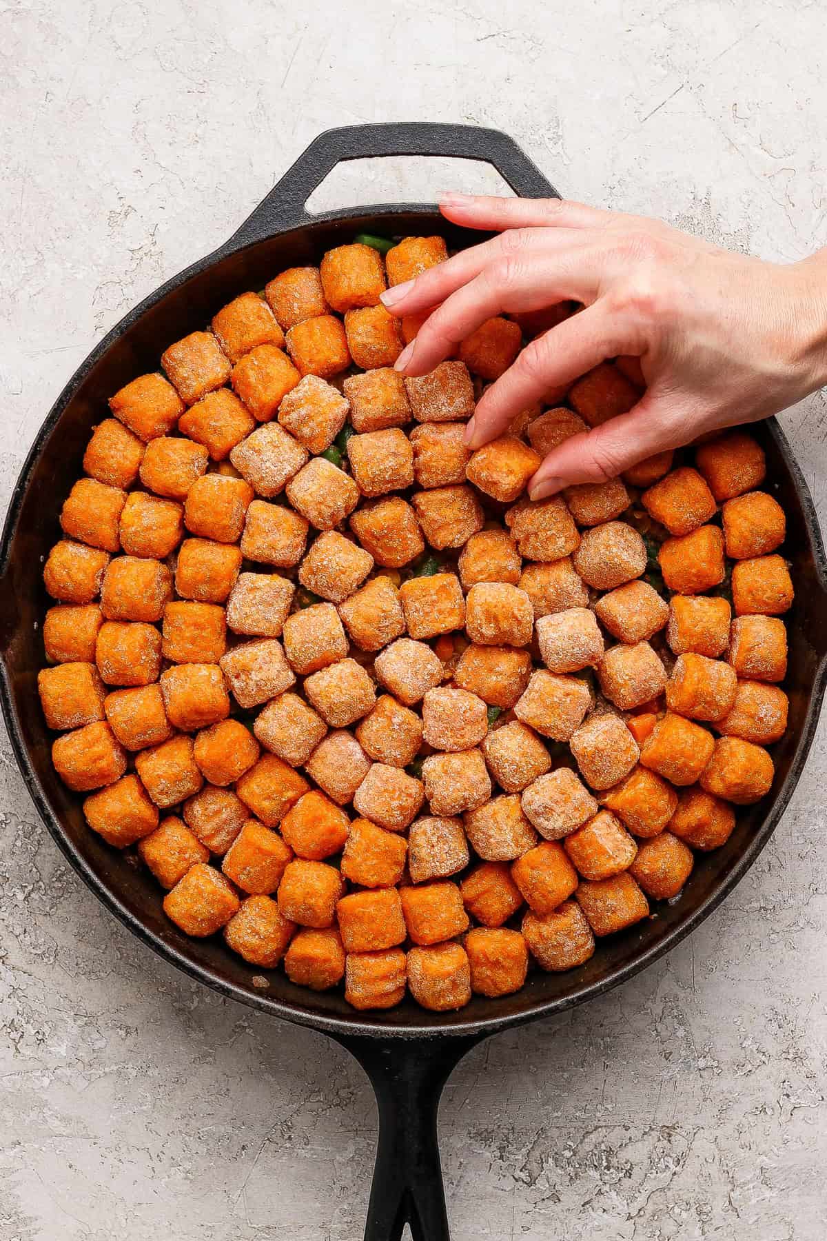 A hand arranges frozen tater tots in a single layer inside a round cast iron skillet on a light textured surface.