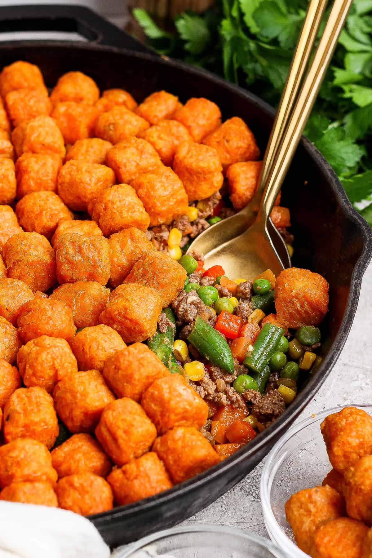 A cast iron skillet filled with ground beef, mixed vegetables, and topped with rows of tater tots. A gold serving spoon is in the skillet.