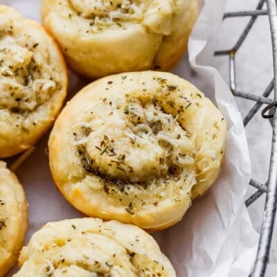 Close-up of savory pastry rolls topped with shredded cheese and herbs, arranged in a metal basket lined with white parchment paper.
