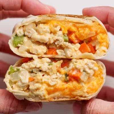 Two hands hold a halved breakfast burrito filled with scrambled eggs, cheese, onions, green peppers, and orange bell peppers.