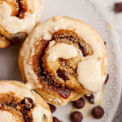 Close-up of cinnamon rolls topped with icing and sprinkled with brown sugar, with chocolate chips scattered on a white plate.
