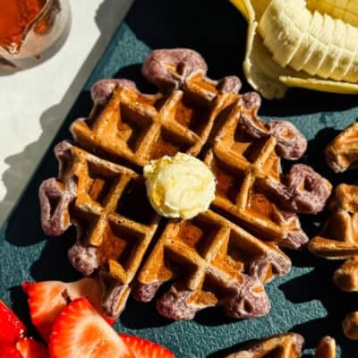 A waffle topped with a dollop of butter is surrounded by sliced bananas and strawberries on a dark surface.
