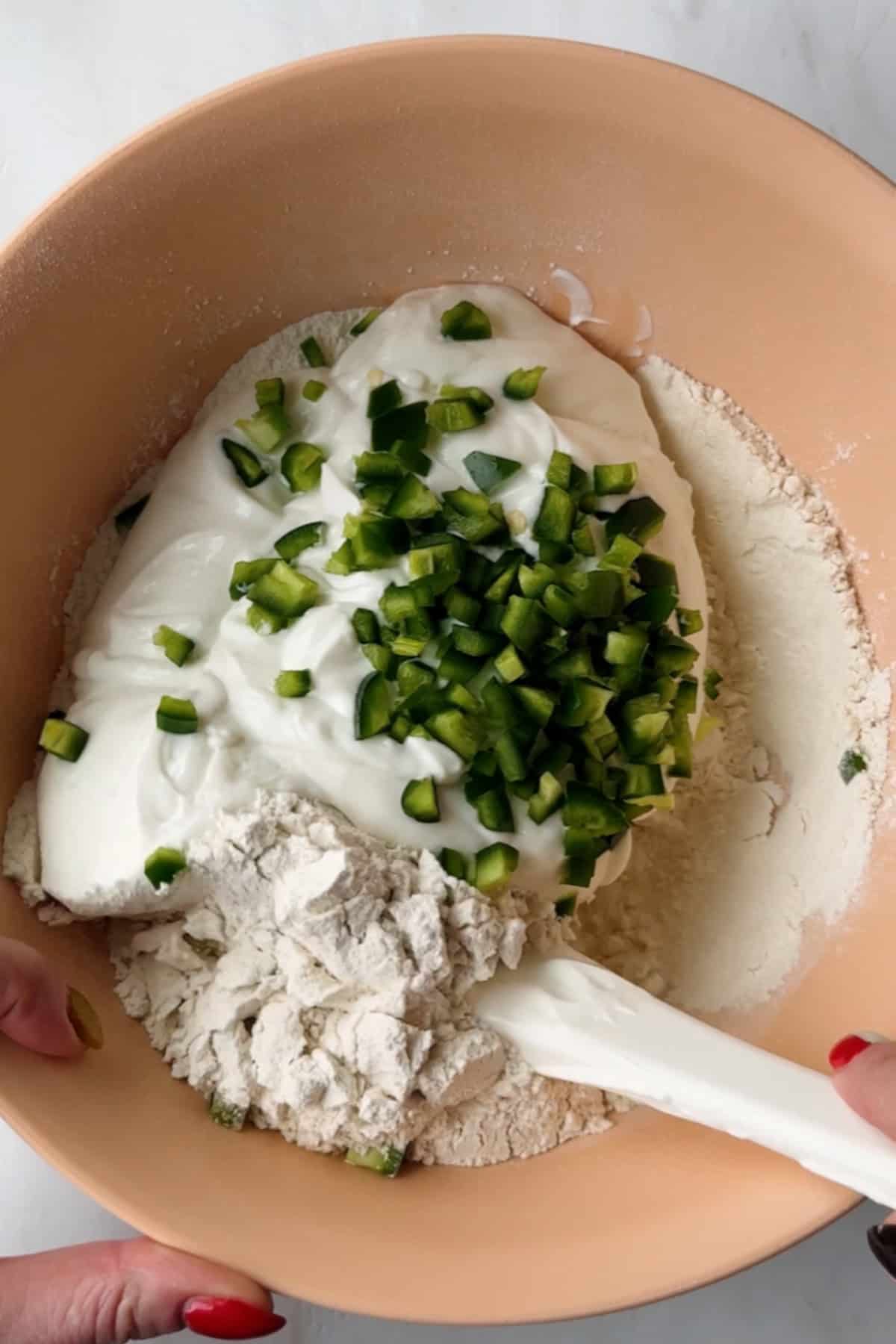 A mixing bowl containing flour, yogurt, and chopped green peppers being stirred with a white spatula.