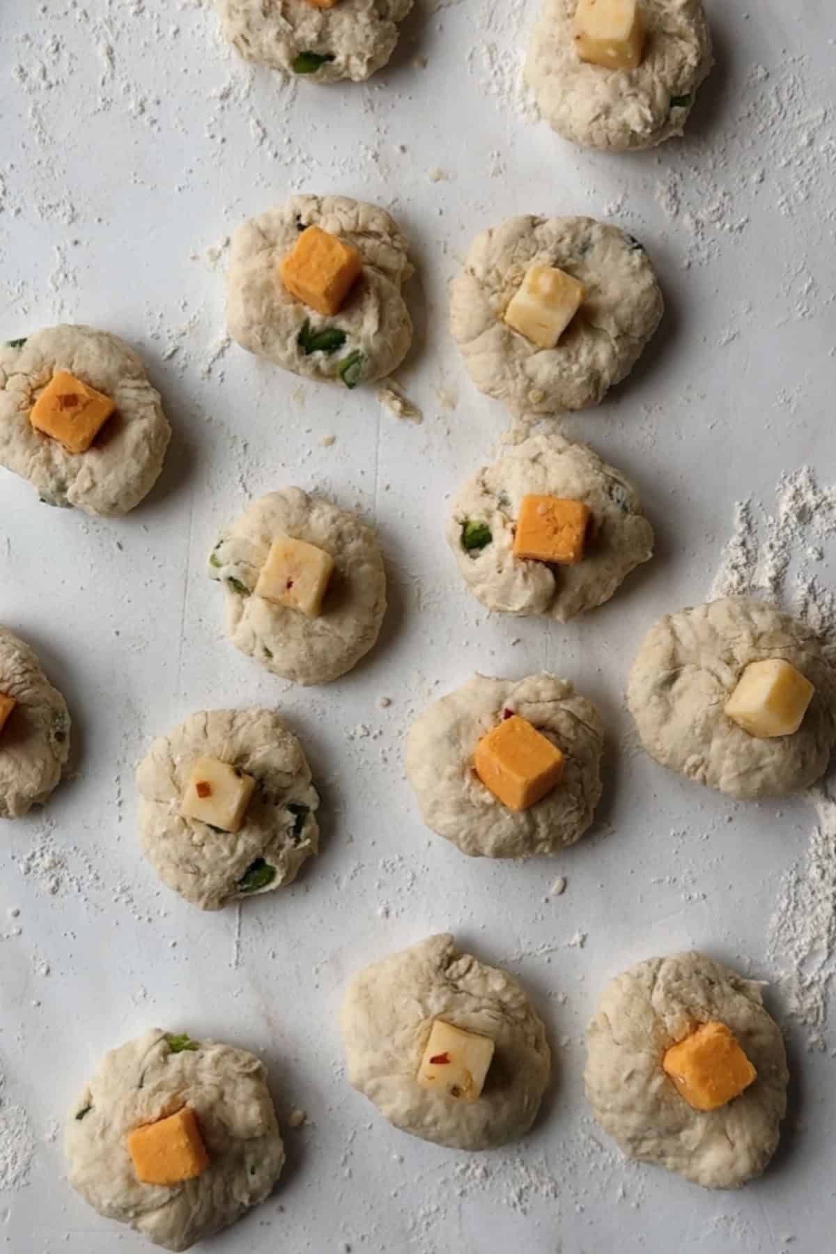 Small rounds of raw dough topped with cubes of cheese are arranged on a lightly floured surface, ready to be baked or cooked.