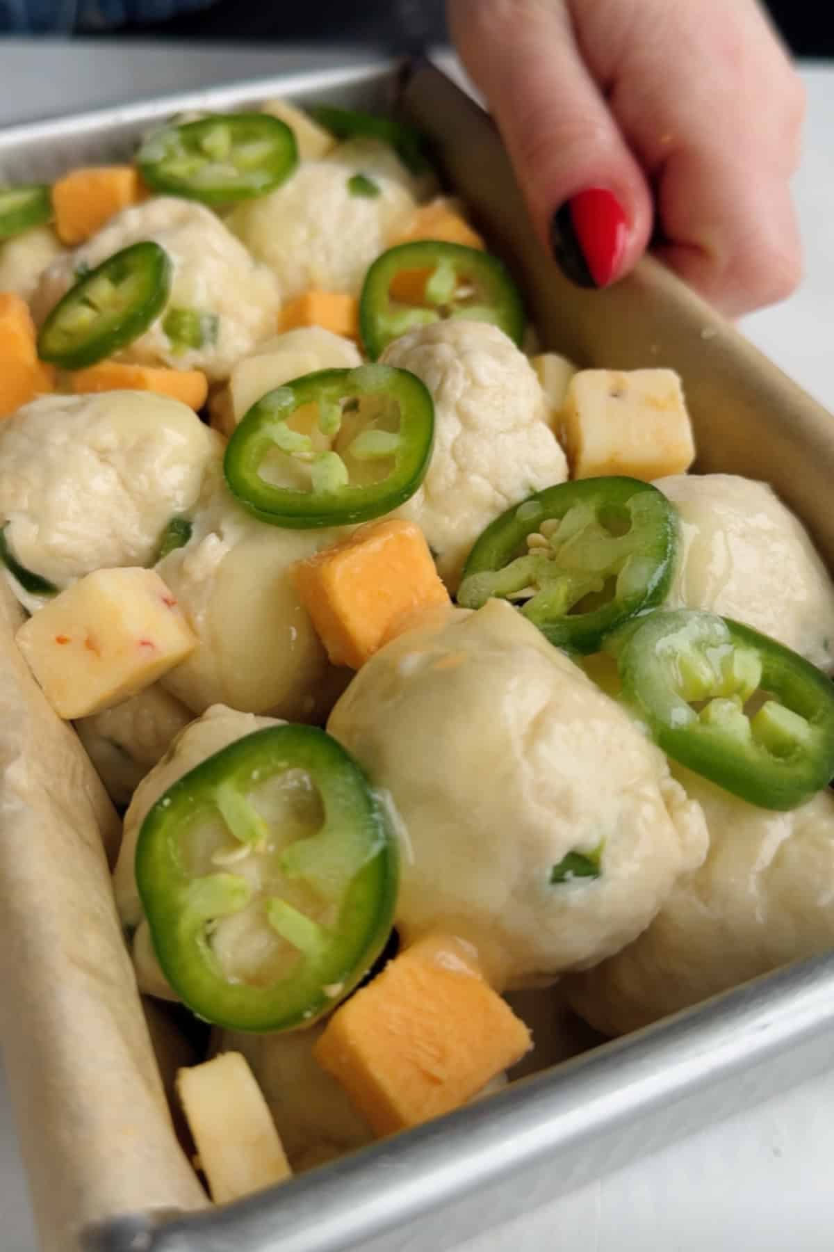 Close-up of unbaked dough balls in a pan, topped with sliced jalape&ntilde;os and cubes of cheddar and pepper jack cheese.