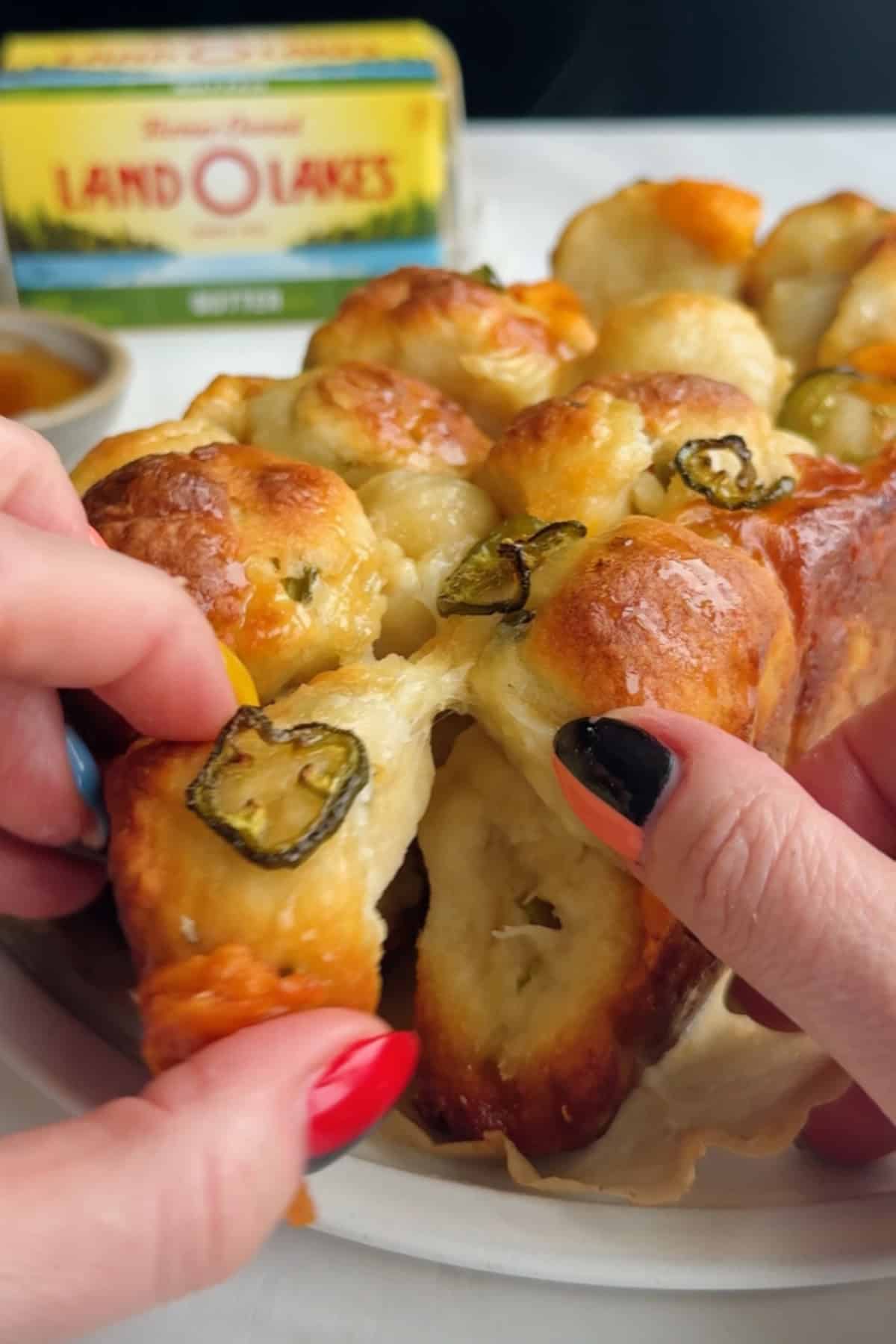 Two hands pull apart a piece of baked bread topped with sliced jalape&ntilde;os, with a package of butter visible in the background.