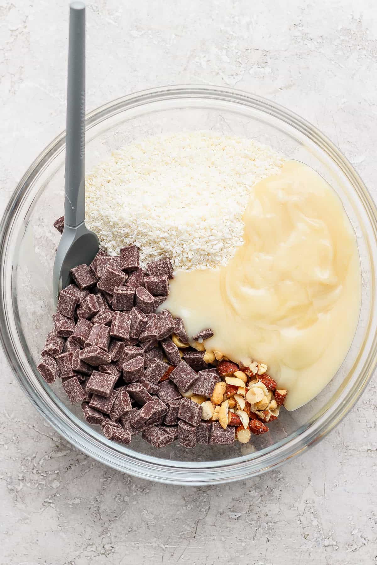 A glass bowl containing sweetened condensed milk, chopped nuts, shredded coconut, and chocolate chunks with a gray spatula.