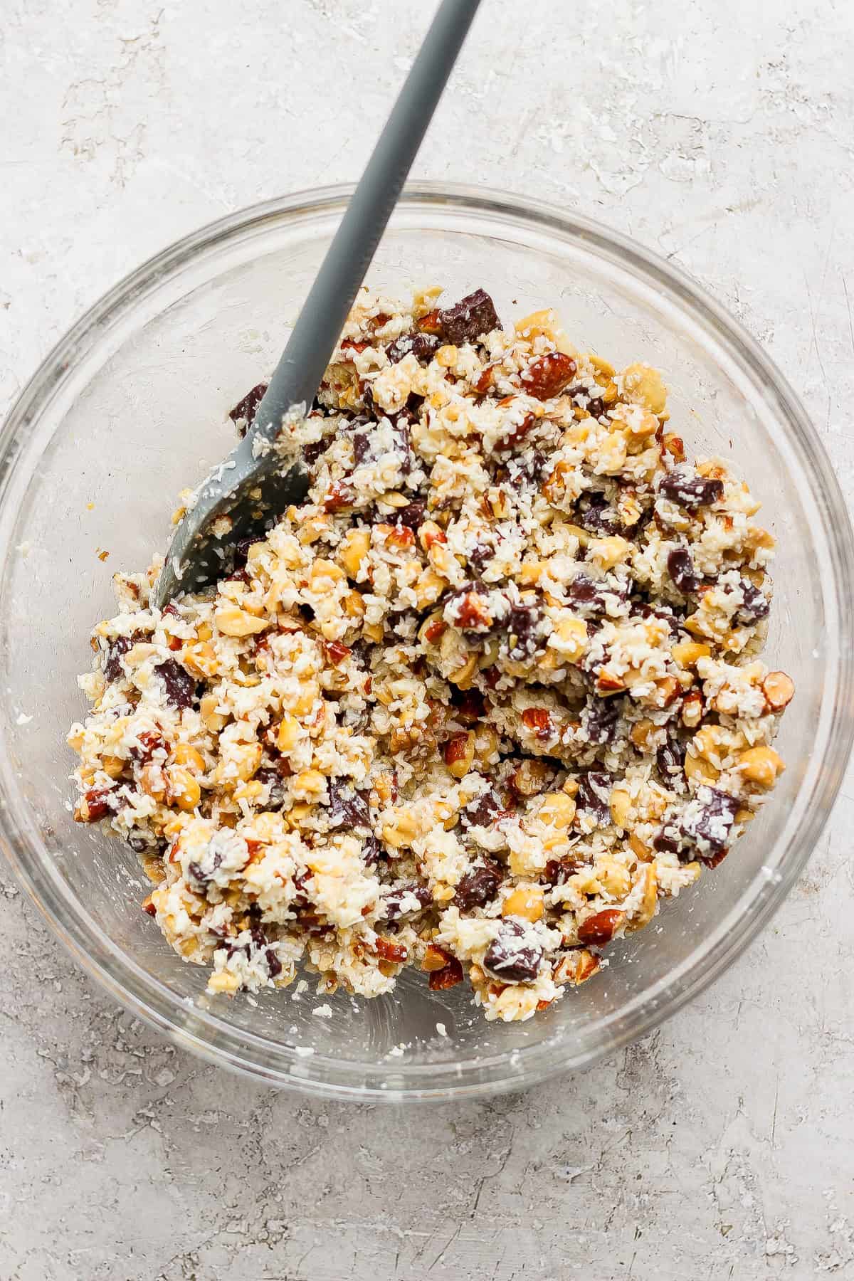 A glass bowl filled with a mixture of chopped nuts, chocolate chunks, and coconut flakes, with a gray spatula resting inside.