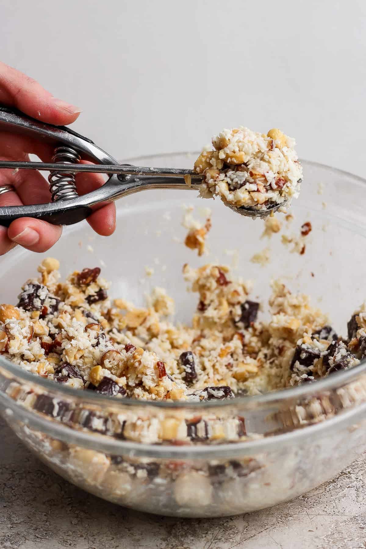 A hand uses a metal cookie scoop to lift dough from a glass bowl filled with a chunky cookie mixture containing nuts and chocolate.