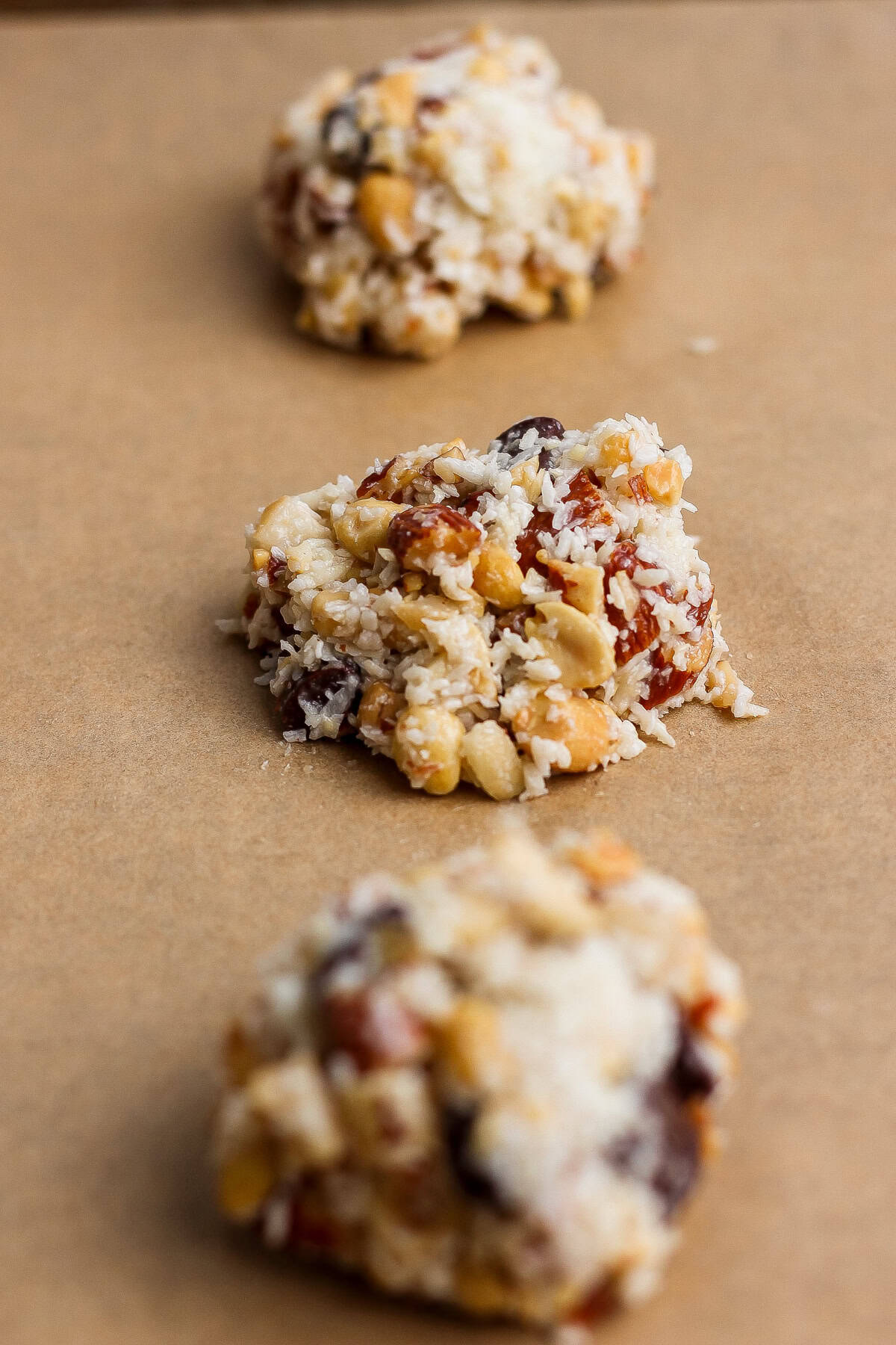 Three unbaked clusters of mixed nuts, dried fruit, and shredded coconut on a sheet of brown parchment paper.