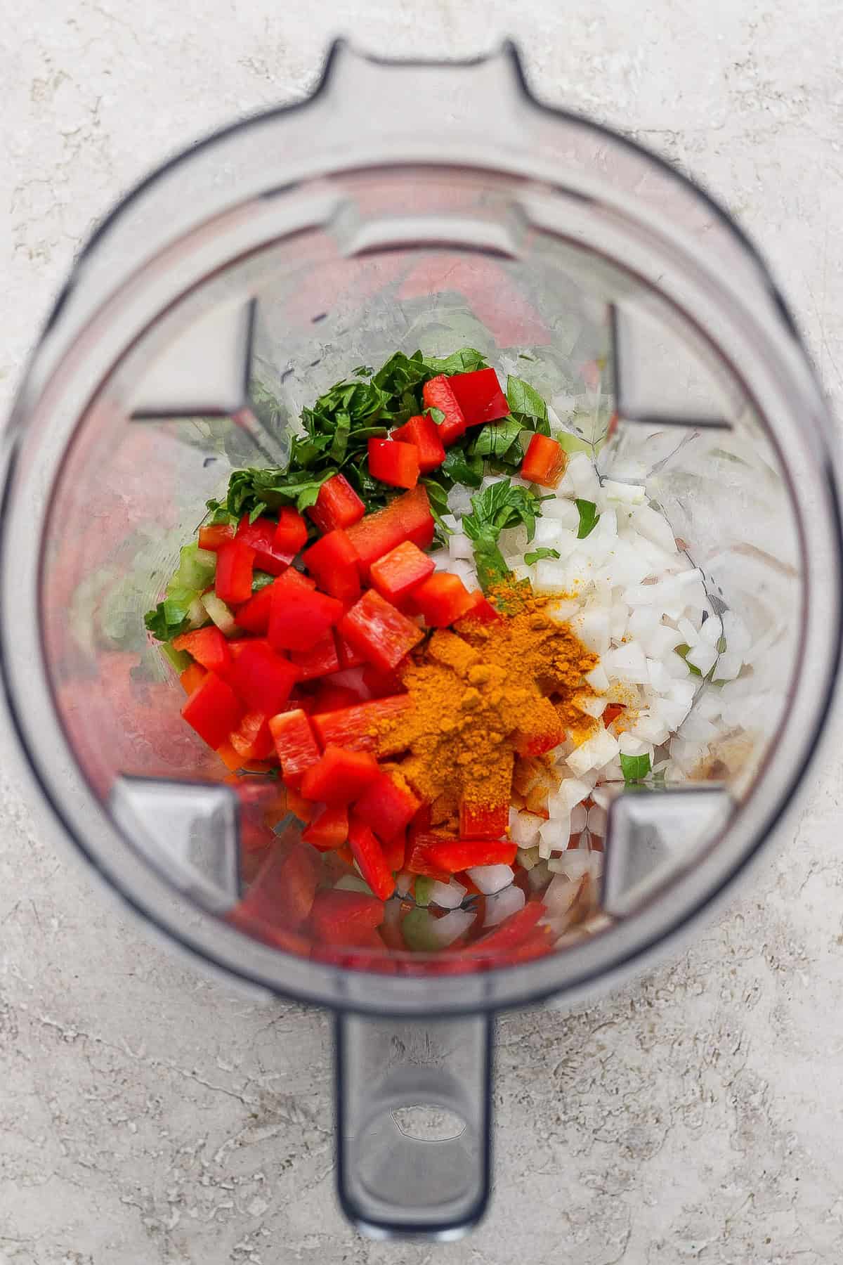 A blender filled with chopped red bell pepper, onion, green herbs, and a bright yellow spice on a light-colored surface.