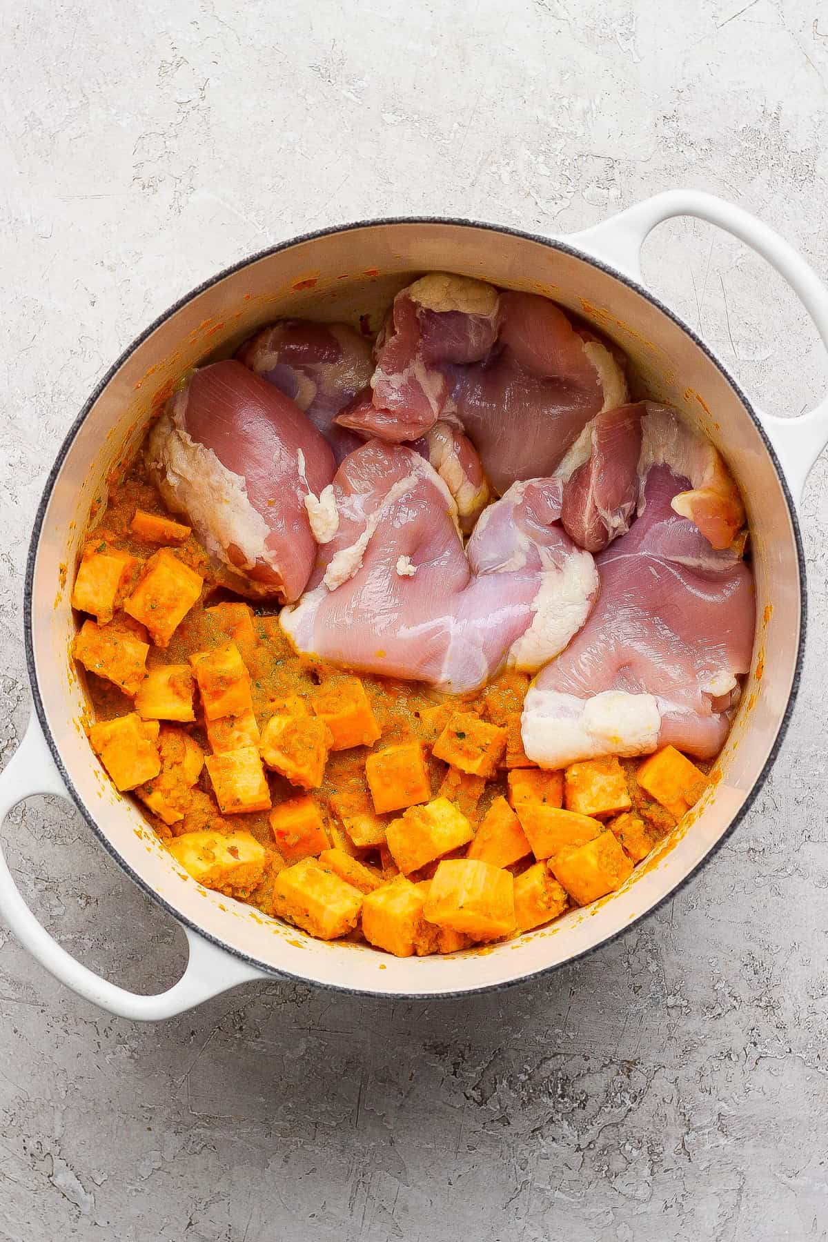 Raw chicken thighs and cubed sweet potatoes in a white Dutch oven on a light textured surface.