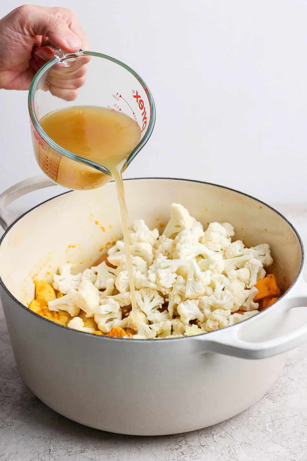 A hand pours broth from a glass measuring cup into a white pot filled with cauliflower florets and other vegetables.
