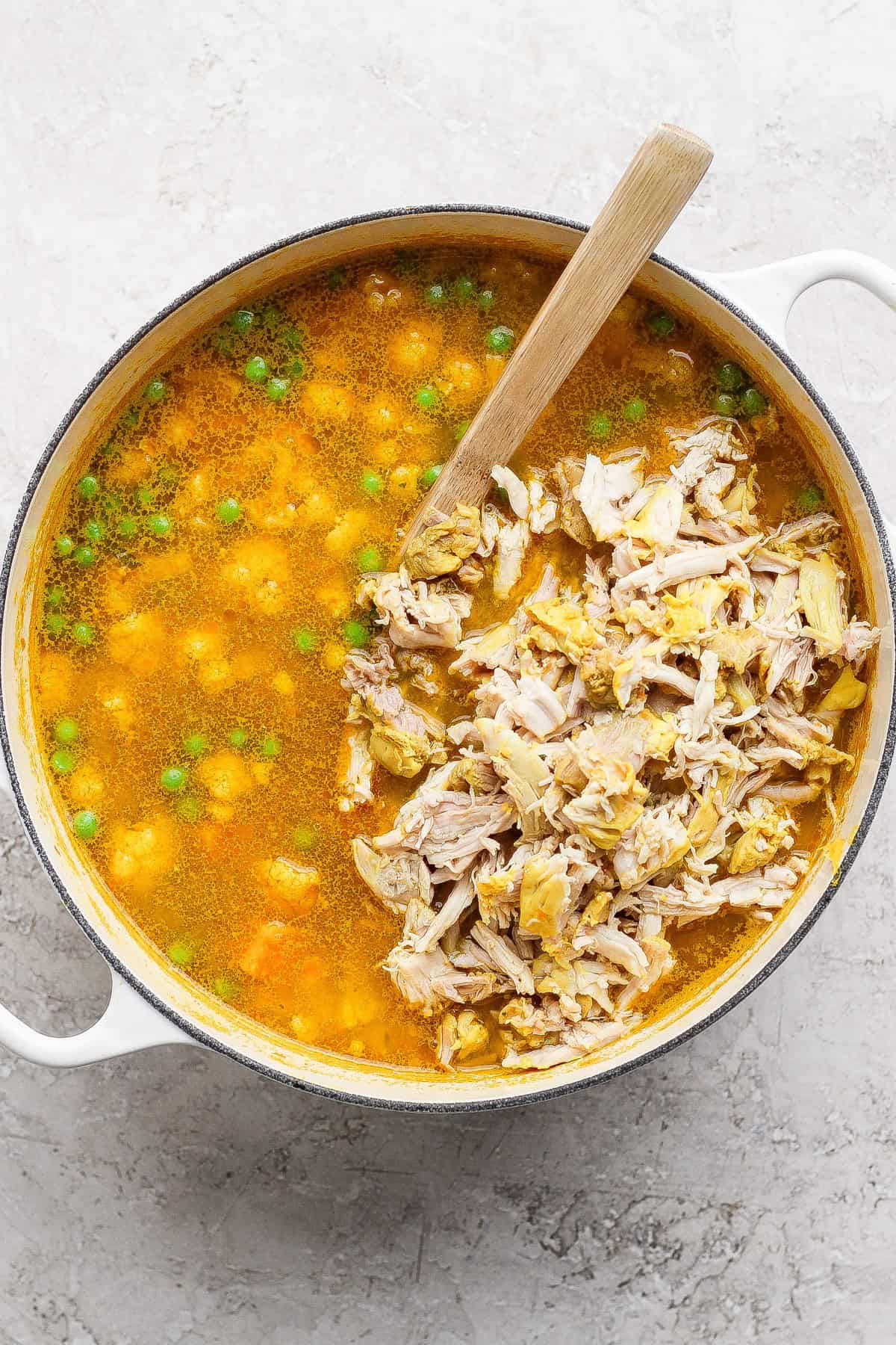 A pot of soup with shredded chicken, green peas, and a wooden spoon on a light-colored surface.