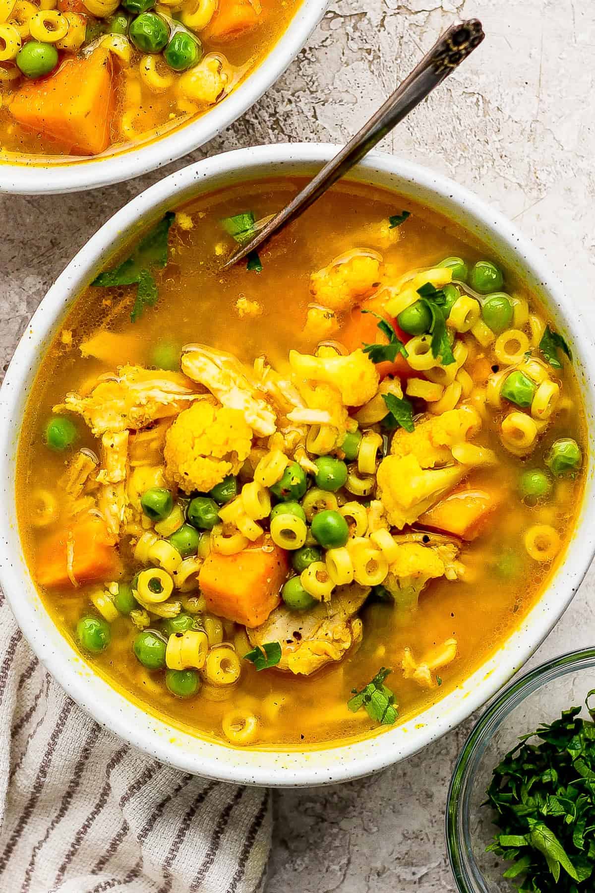 A bowl of chicken noodle soup with round pasta, peas, cauliflower, carrots, and herbs, with a spoon and a striped cloth nearby.
