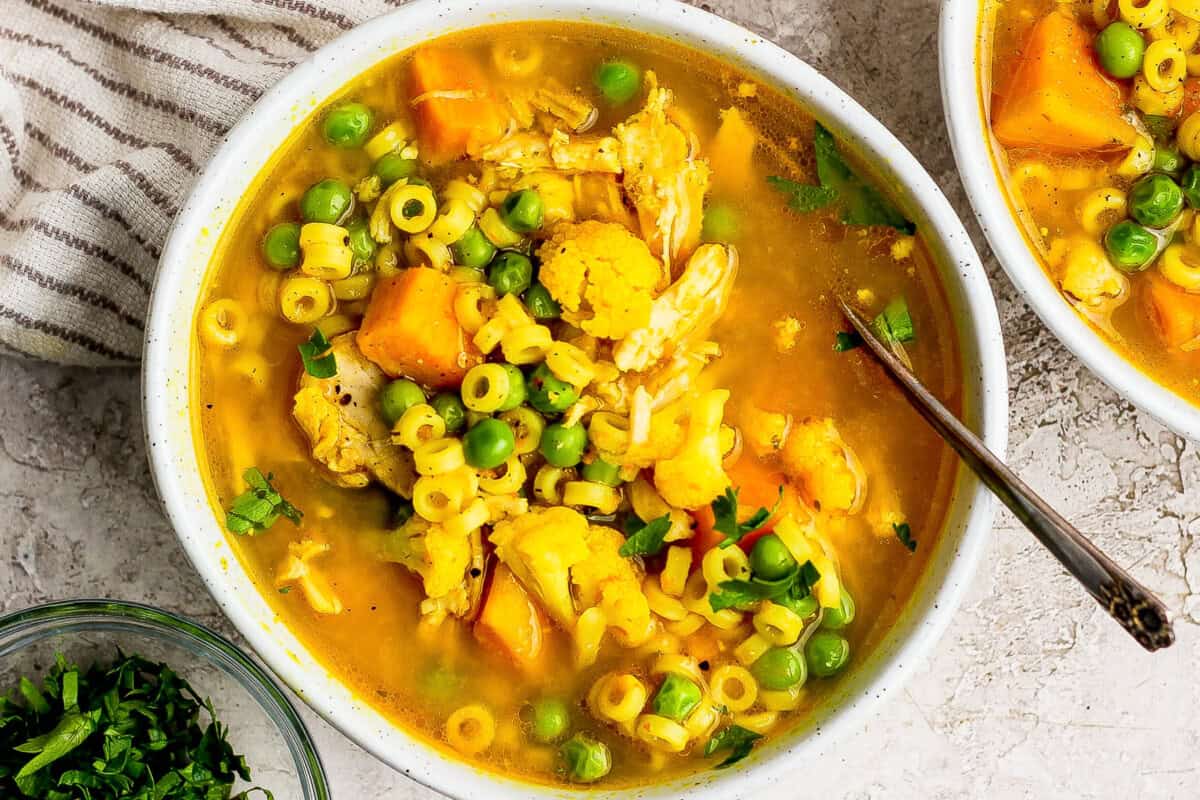 A bowl of yellow soup with small pasta, peas, cauliflower, and chunks of orange vegetables, garnished with chopped herbs, and a spoon inside the bowl.