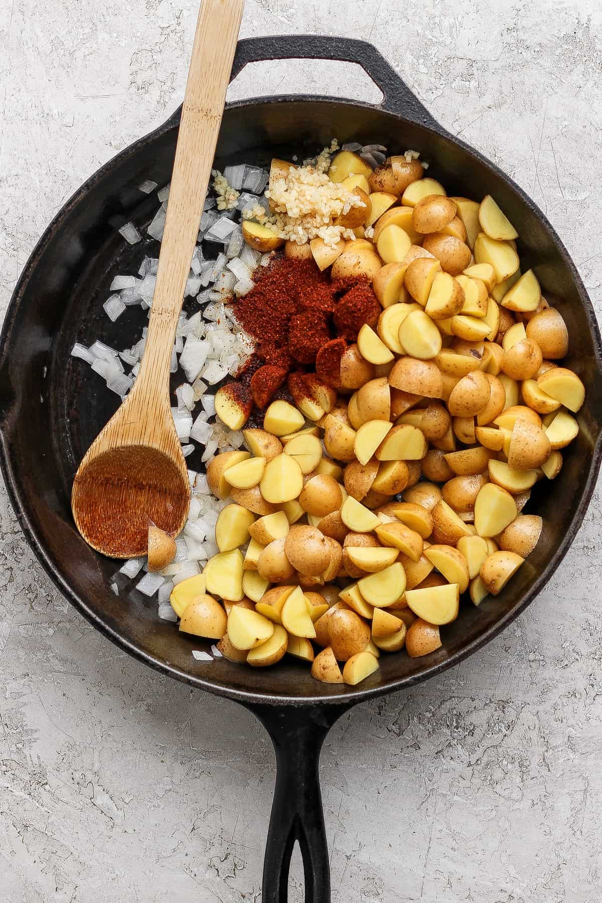 A cast iron skillet filled with chopped potatoes, diced onions, minced garlic, and paprika, with a wooden spoon resting on the side.