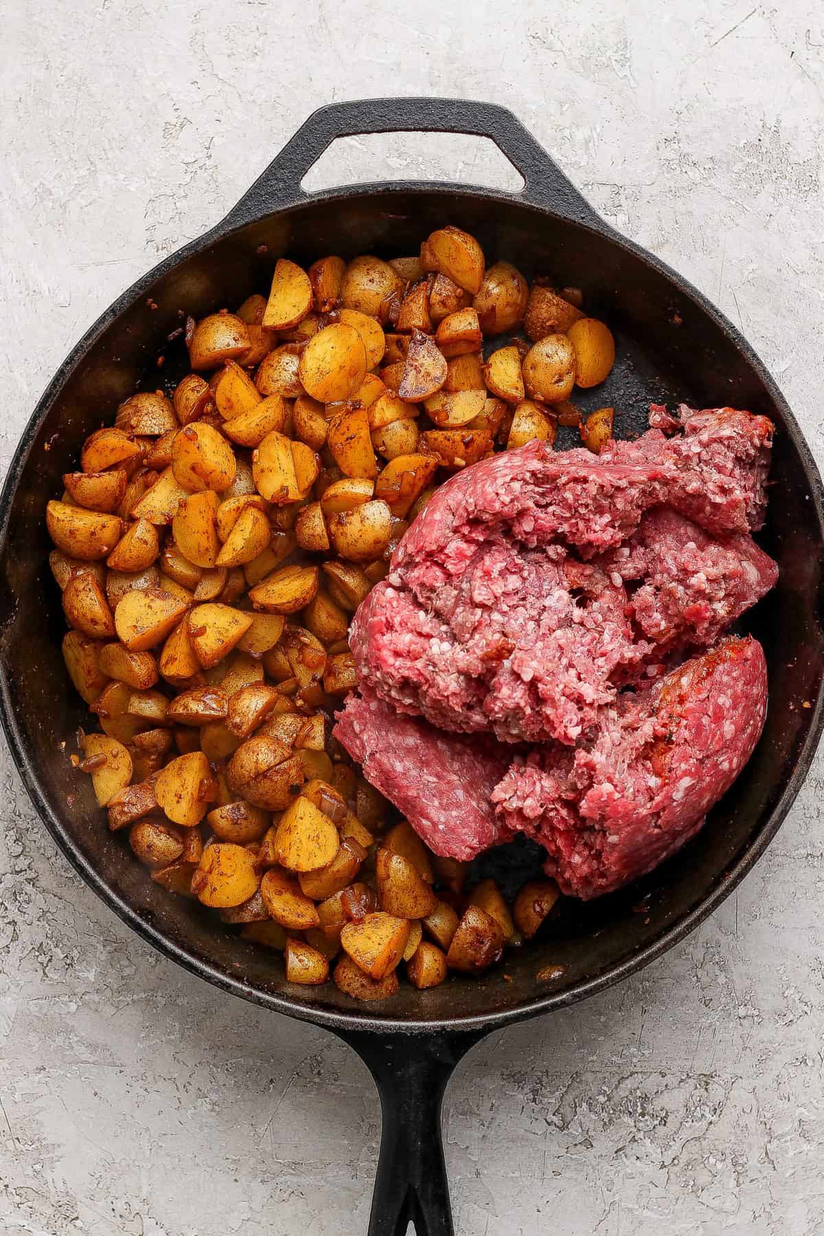 A cast iron skillet with cooked diced potatoes on one side and raw ground beef on the other, on a light textured surface.