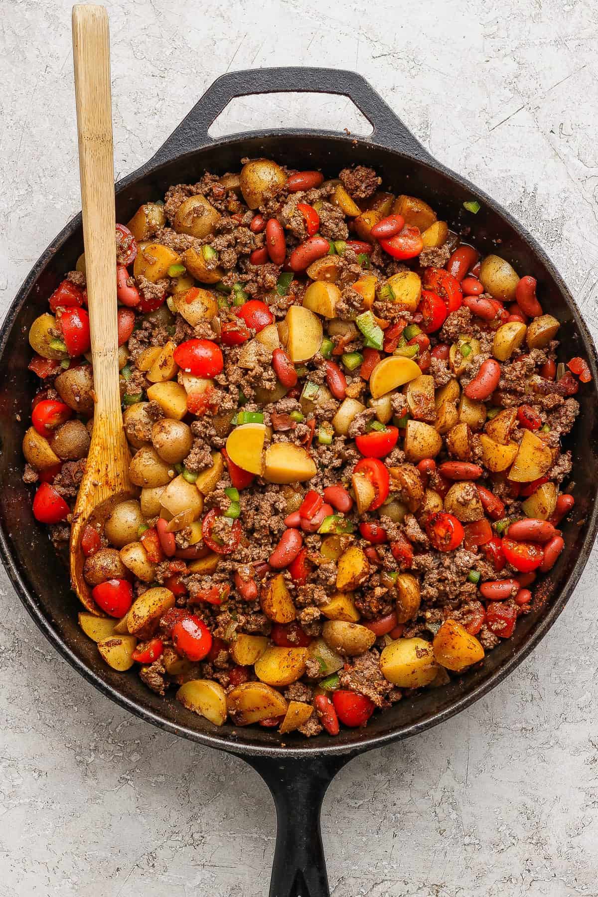 A cast iron skillet filled with cooked ground beef, halved baby potatoes, kidney beans, cherry tomatoes, and chopped green onions, with a wooden spoon resting inside.