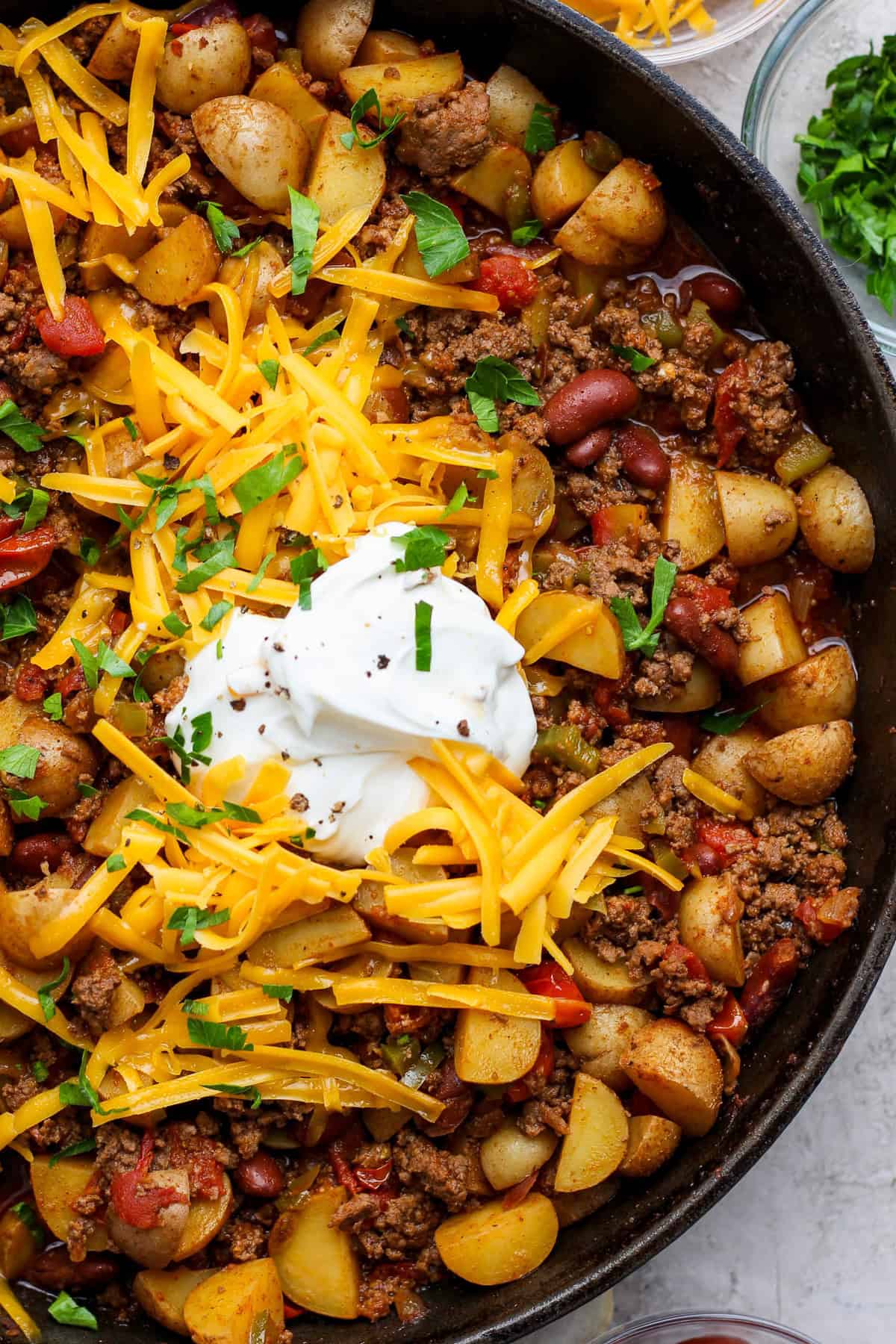A skillet filled with ground beef, kidney beans, potatoes, and tomatoes, topped with shredded cheddar cheese, sour cream, and chopped parsley.