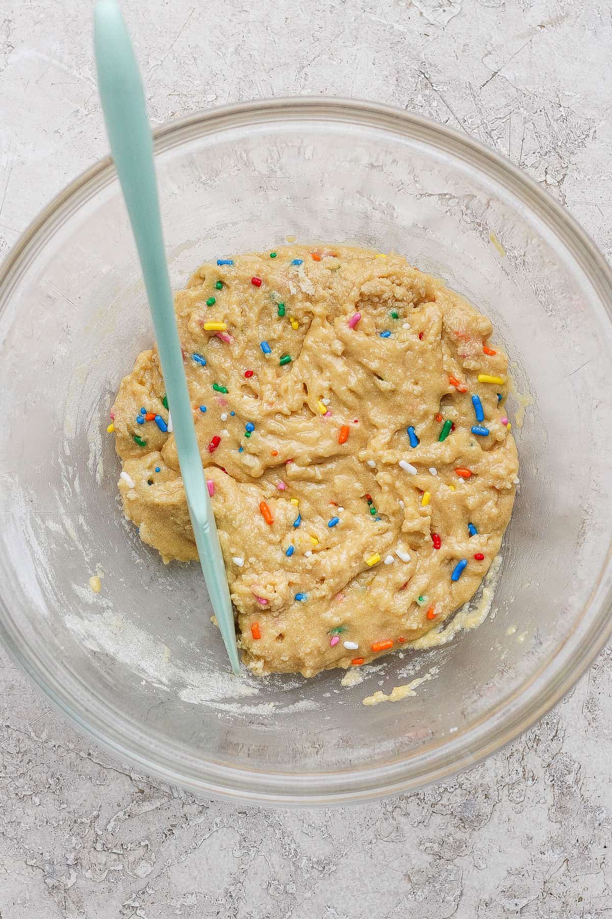A glass bowl containing raw cookie dough with colorful sprinkles and a light blue spatula on a textured surface.