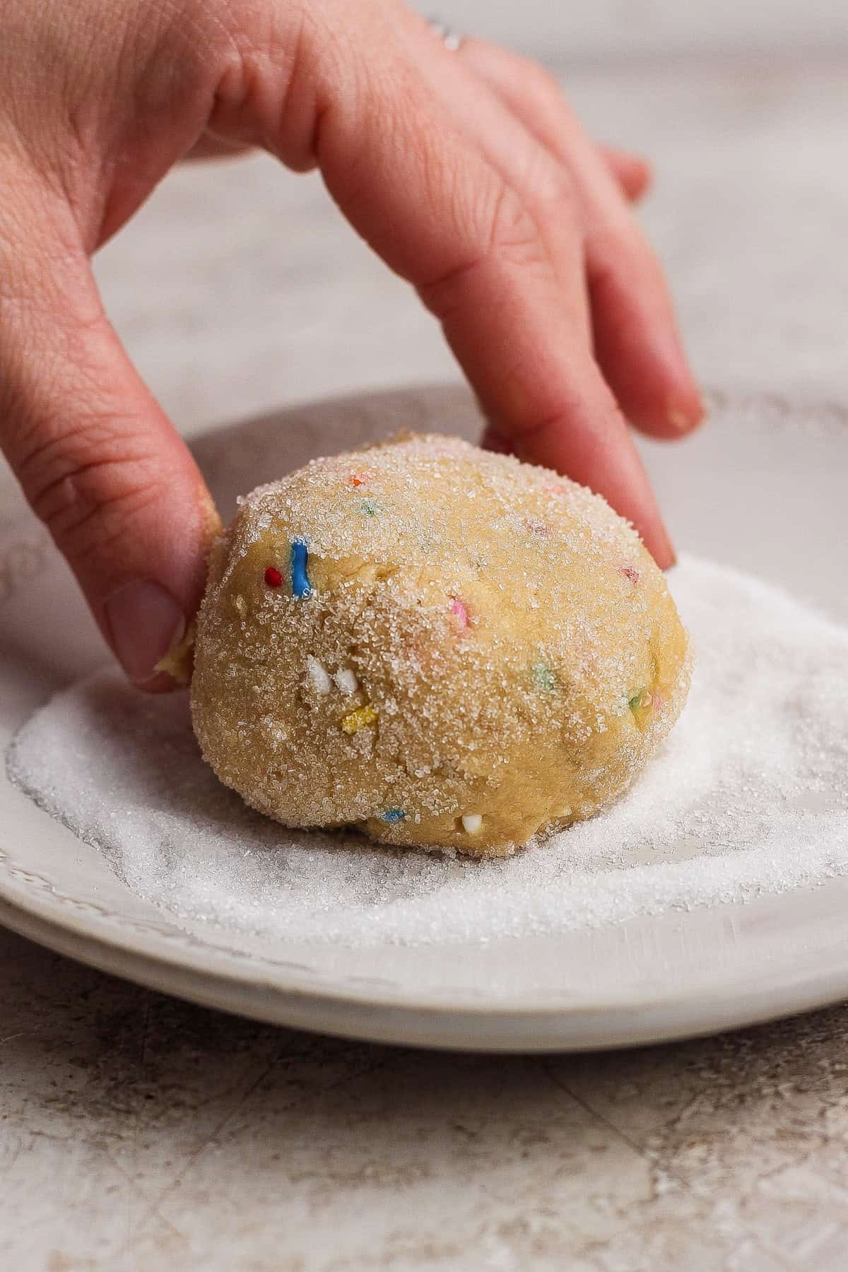 A hand rolls a ball of cookie dough with colorful sprinkles in a layer of white sugar on a plate.