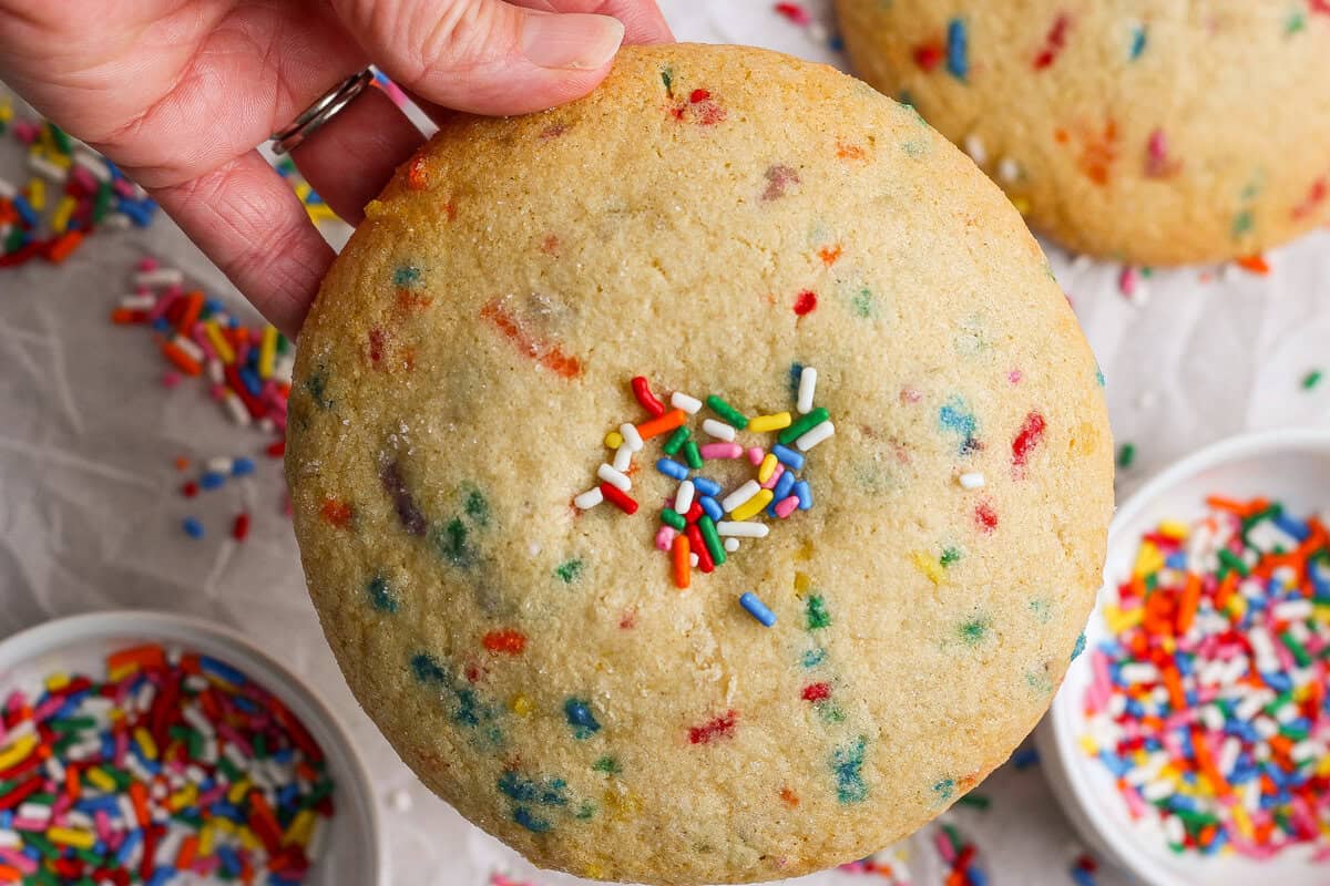 A hand holding a large sugar cookie with colorful sprinkles, surrounded by bowls of sprinkles on a light surface.