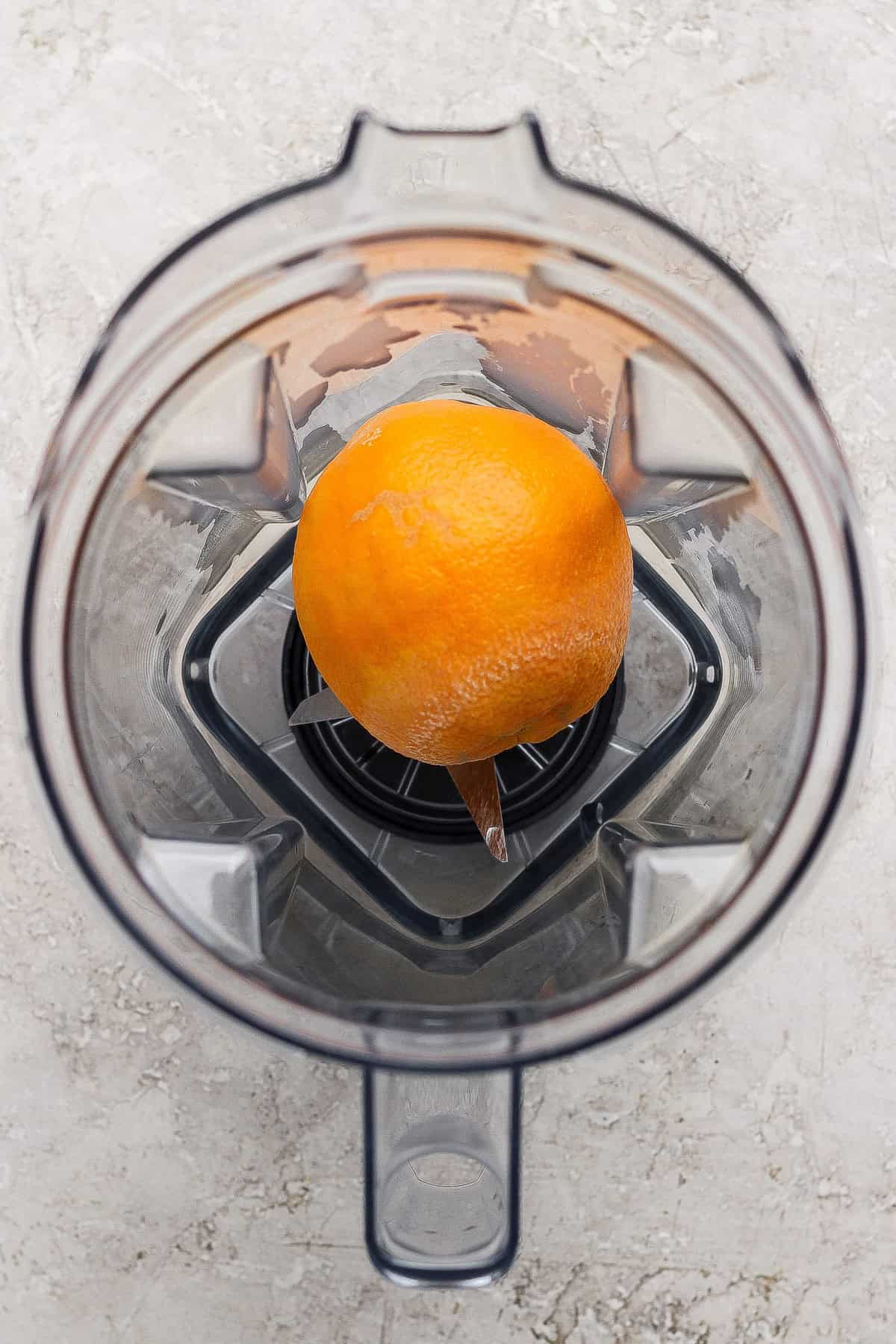 A whole orange with a small peel removed sits at the bottom of an empty blender viewed from above.