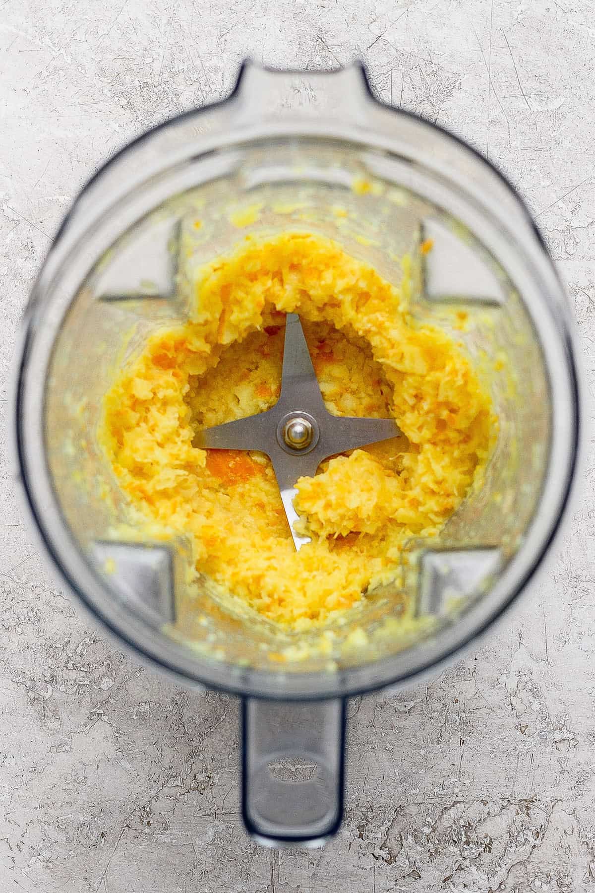 Top-down view of a blender with a yellow-orange mixture partially pureed, showing metal blades and textured sides, on a light gray surface.
