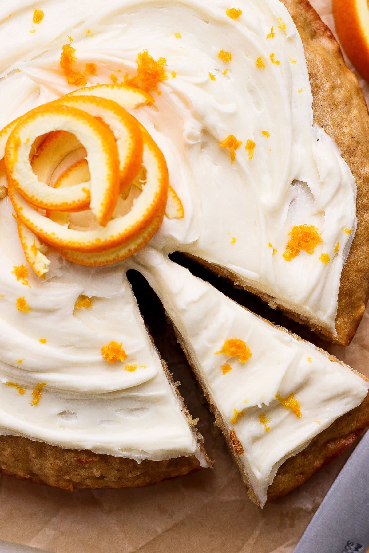A round cake with white frosting, garnished with orange zest and a spiral of orange peel, with one slice being separated from the whole cake.