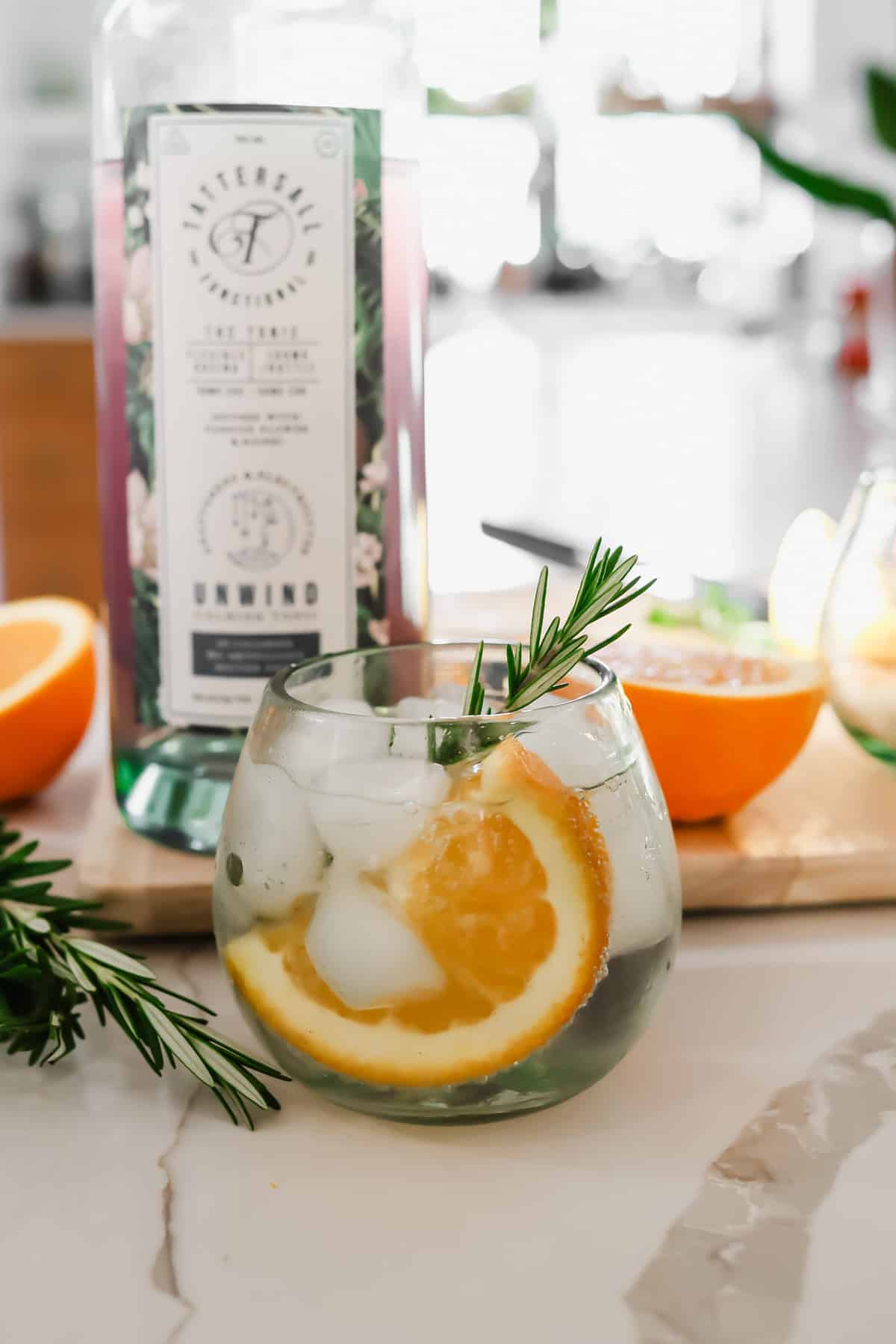 A glass of iced drink garnished with orange slices and rosemary sits in front of a bottle labeled "Unwind" and a halved orange on a countertop.