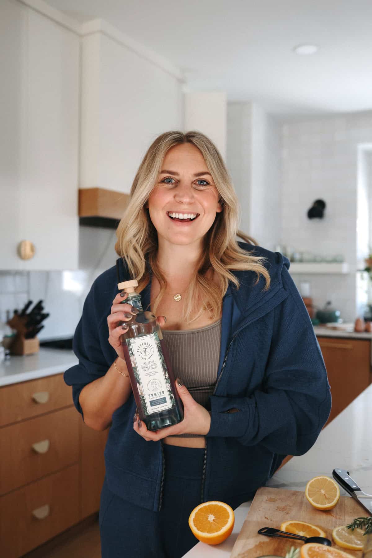 A woman in a blue tracksuit smiles while holding a bottle in a kitchen with oranges sliced on a cutting board in front of her.