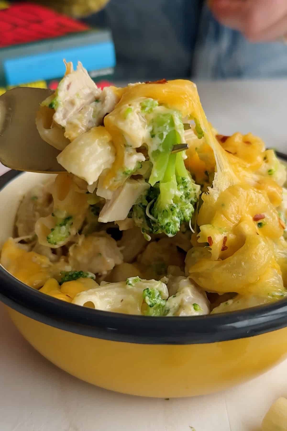 A fork lifts a portion of cheesy pasta with broccoli and chicken from a yellow enamel bowl.