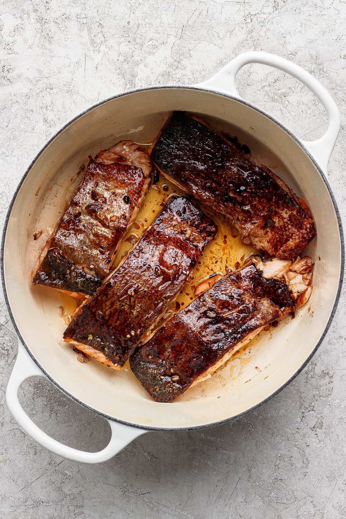 Four seared salmon fillets with browned skin cooking in a white enameled cast iron pan on a light textured surface.