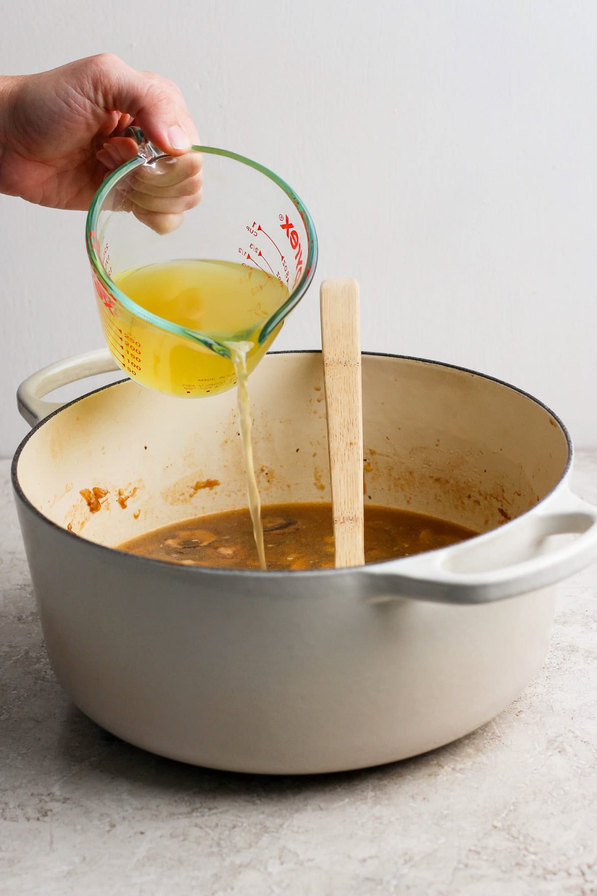 A hand pours broth from a measuring cup into a large pot containing a brown mixture, with a wooden spatula resting inside the pot.