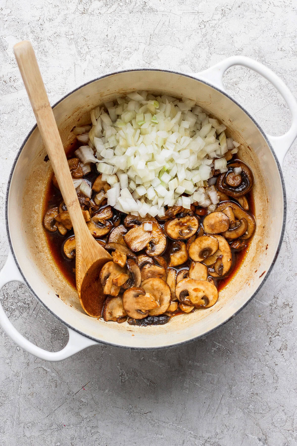 A white pot containing sliced mushrooms in sauce and a pile of diced onions, with a wooden spoon resting inside.