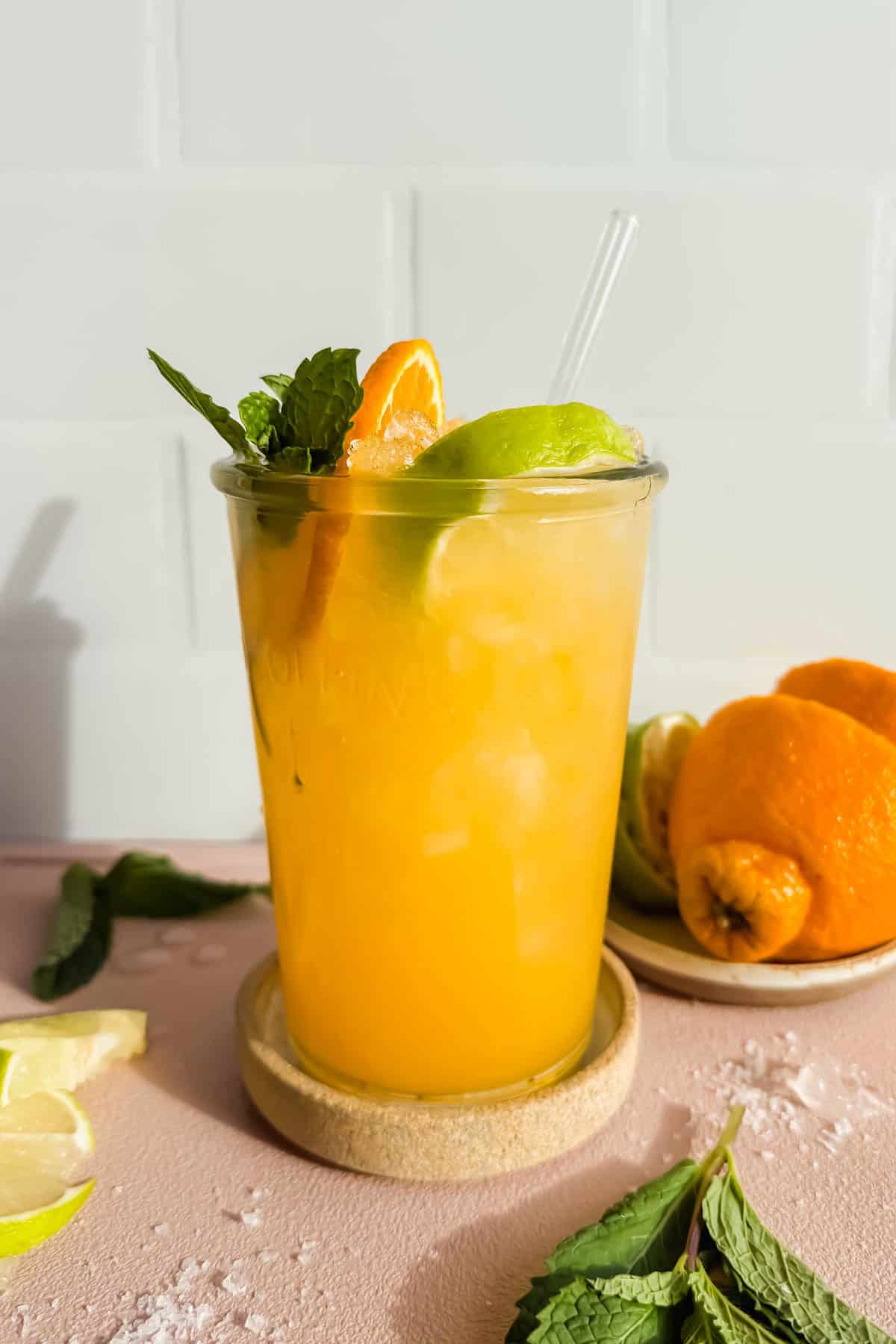 A glass of orange-colored iced drink garnished with lime, orange slice, and mint, with a straw, sits on a coaster next to fresh citrus fruits and mint leaves.