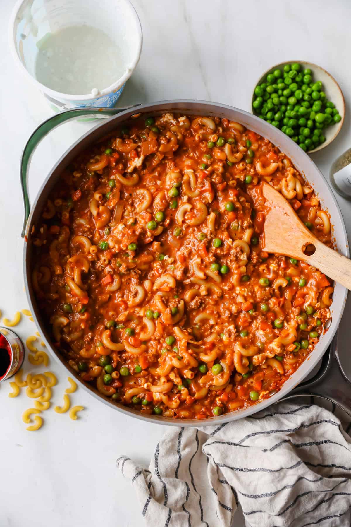A pan of macaroni in tomato sauce with peas is being stirred with a wooden spoon. A bowl of peas, some dry macaroni, and a striped towel are nearby on a white surface.