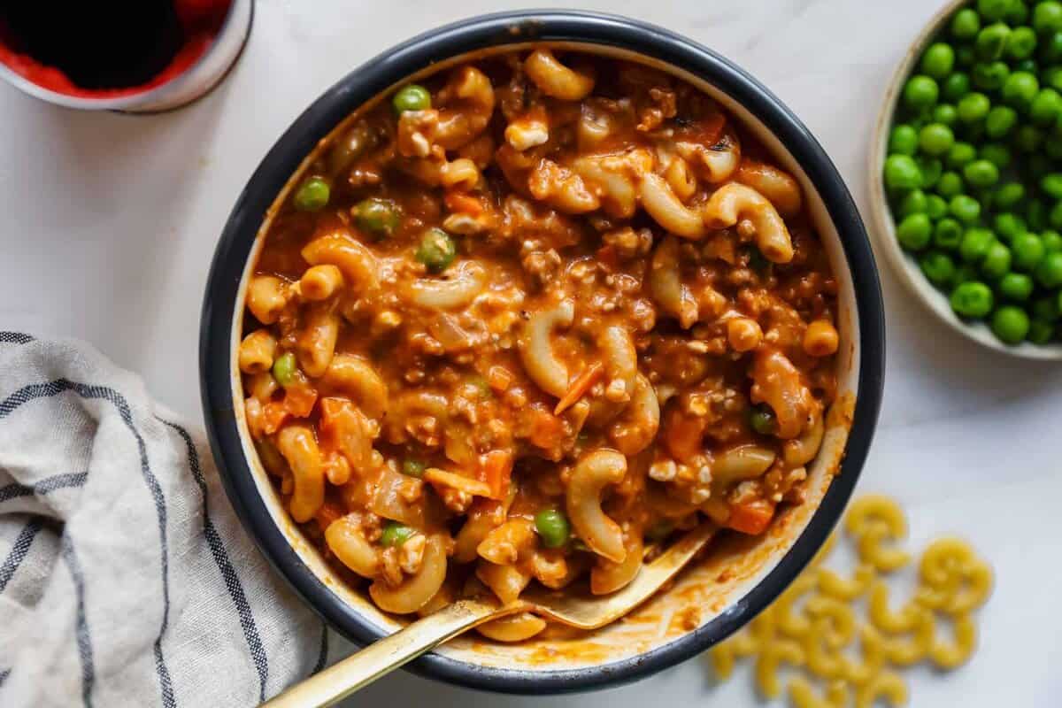 A bowl of macaroni in a thick tomato-based sauce with ground meat, peas, and carrots, with a spoon inside. A bowl of green peas and some dry macaroni are nearby.