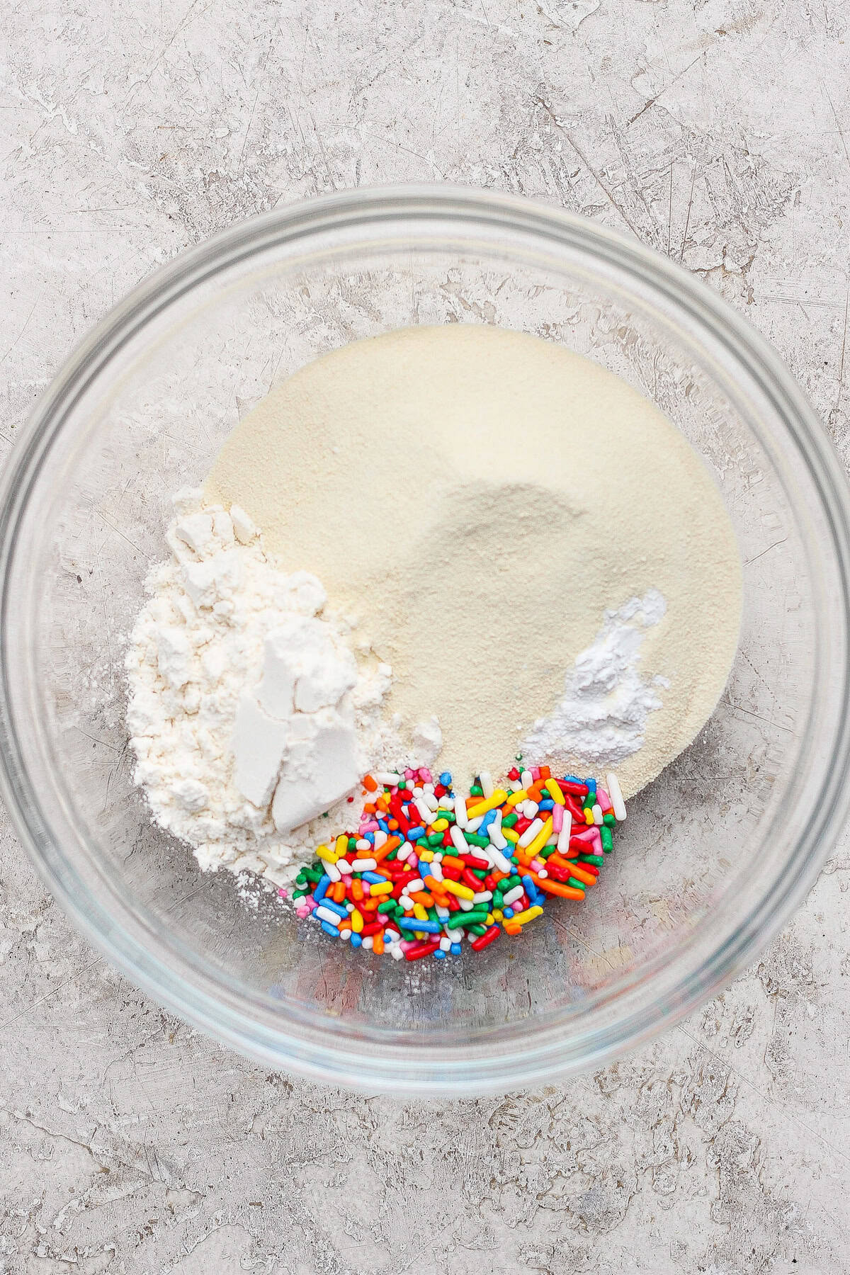 A glass bowl containing white flour, granulated sugar, baking powder, and colorful rainbow sprinkles on a light countertop.