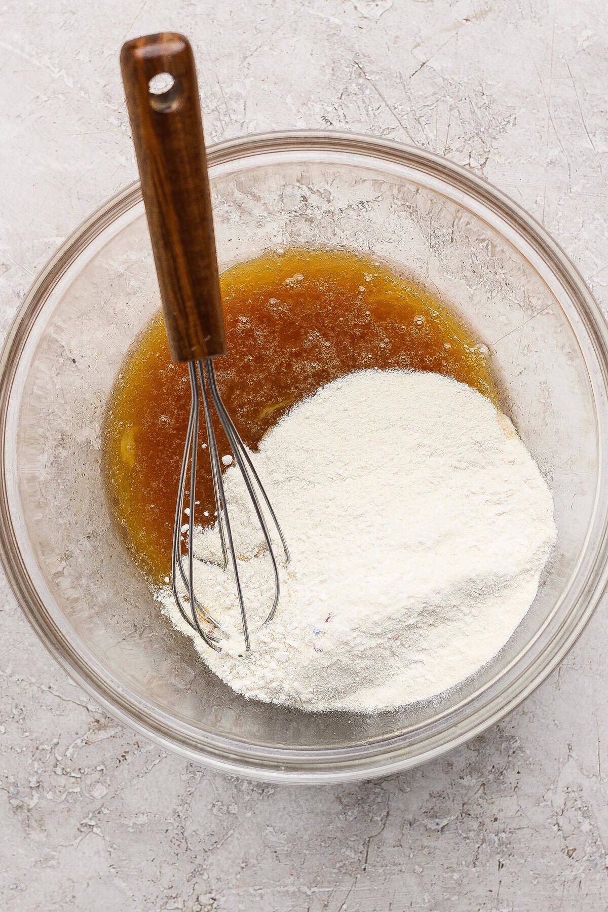 A glass bowl containing flour and a brown liquid being mixed with a metal whisk on a light textured surface.