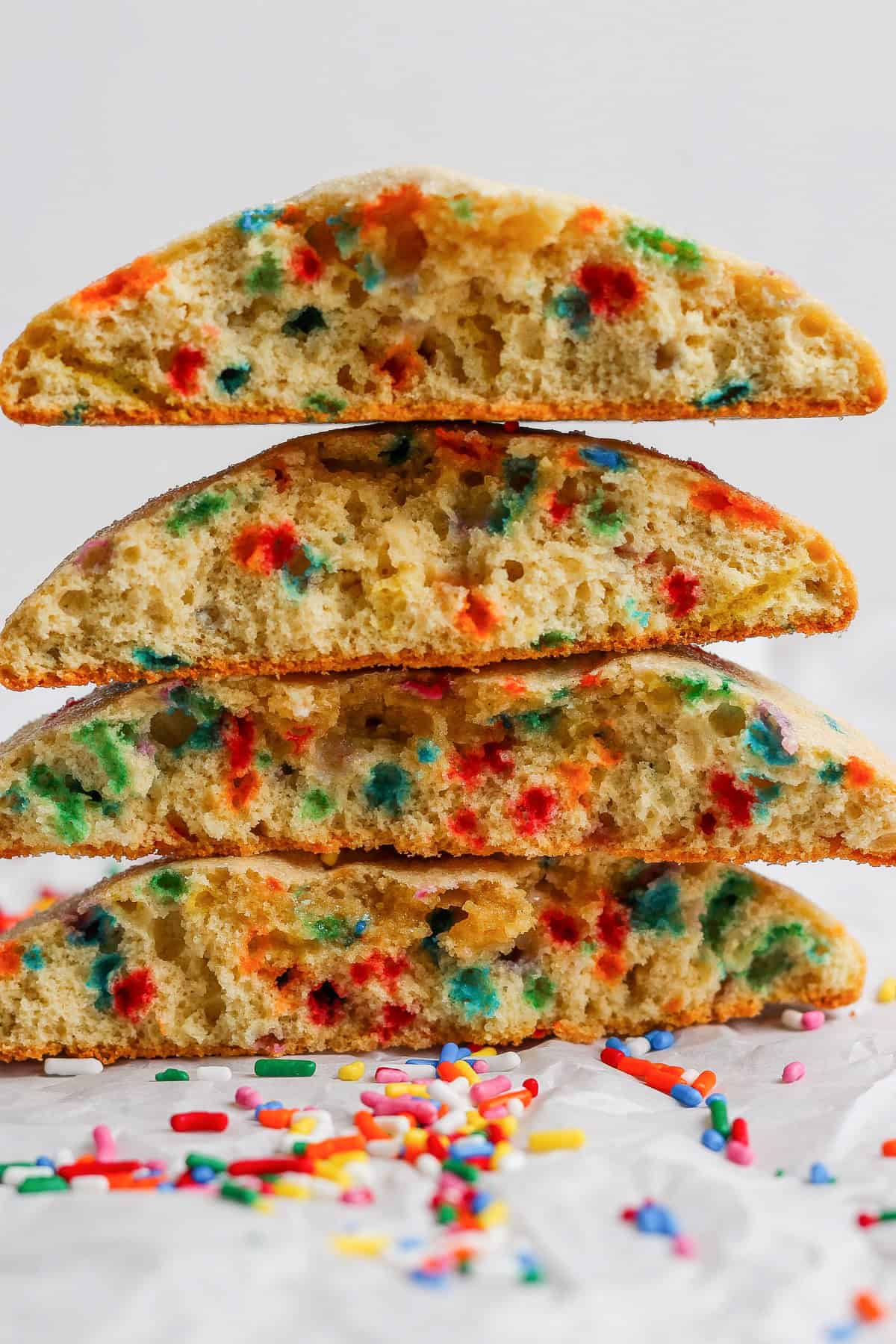 Four thick, halved cookies with colorful sprinkles inside are stacked on parchment paper, with extra sprinkles scattered below.
