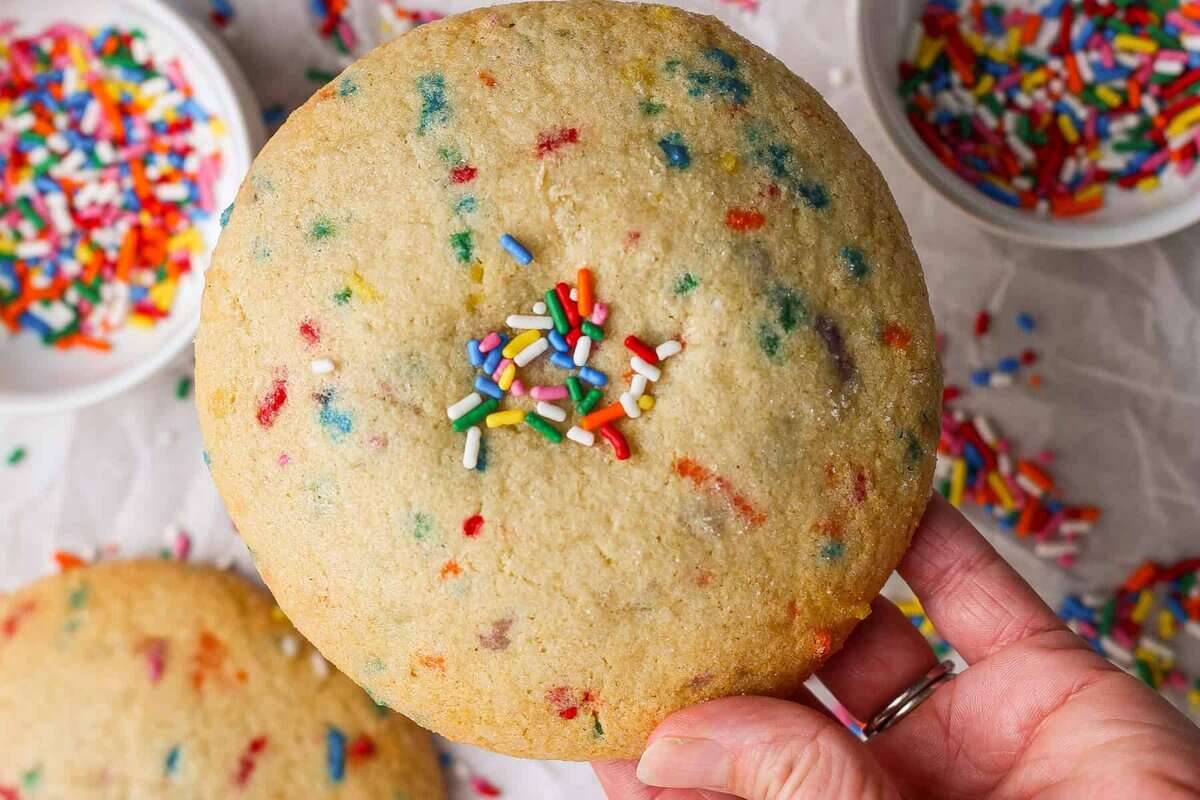 A hand holds a large sugar cookie with colorful sprinkles, with bowls of additional sprinkles and another cookie in the background.