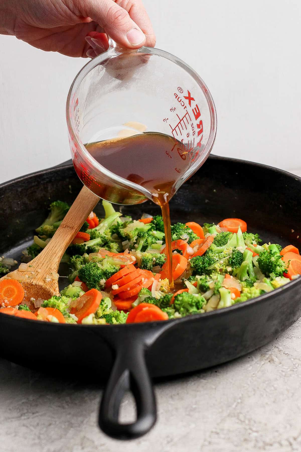 A hand pours sauce from a measuring cup into a cast iron skillet with broccoli and sliced carrots being stirred with a wooden spoon.